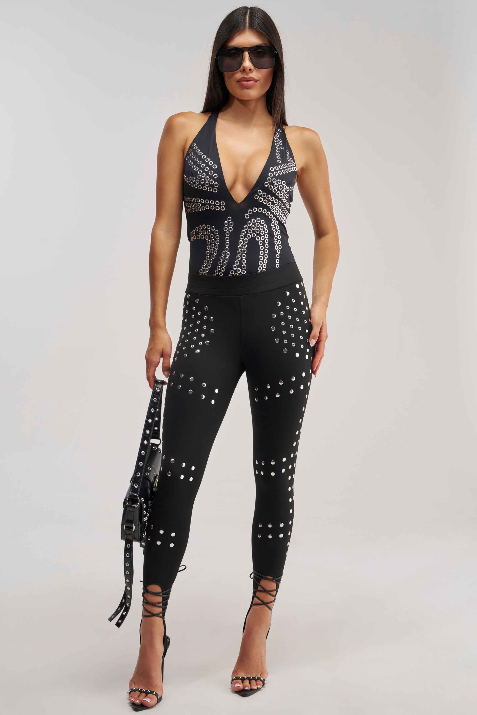 A woman in a black studded outfit with a plunging neckline stands against a plain light background, holding a black handbag. She accessorizes with strappy high heels and sunglasses, completing her chic ensemble with the SNATCHED AND SCULPTED STUD LEGGING that adds an edgy twist to this sophisticated look.