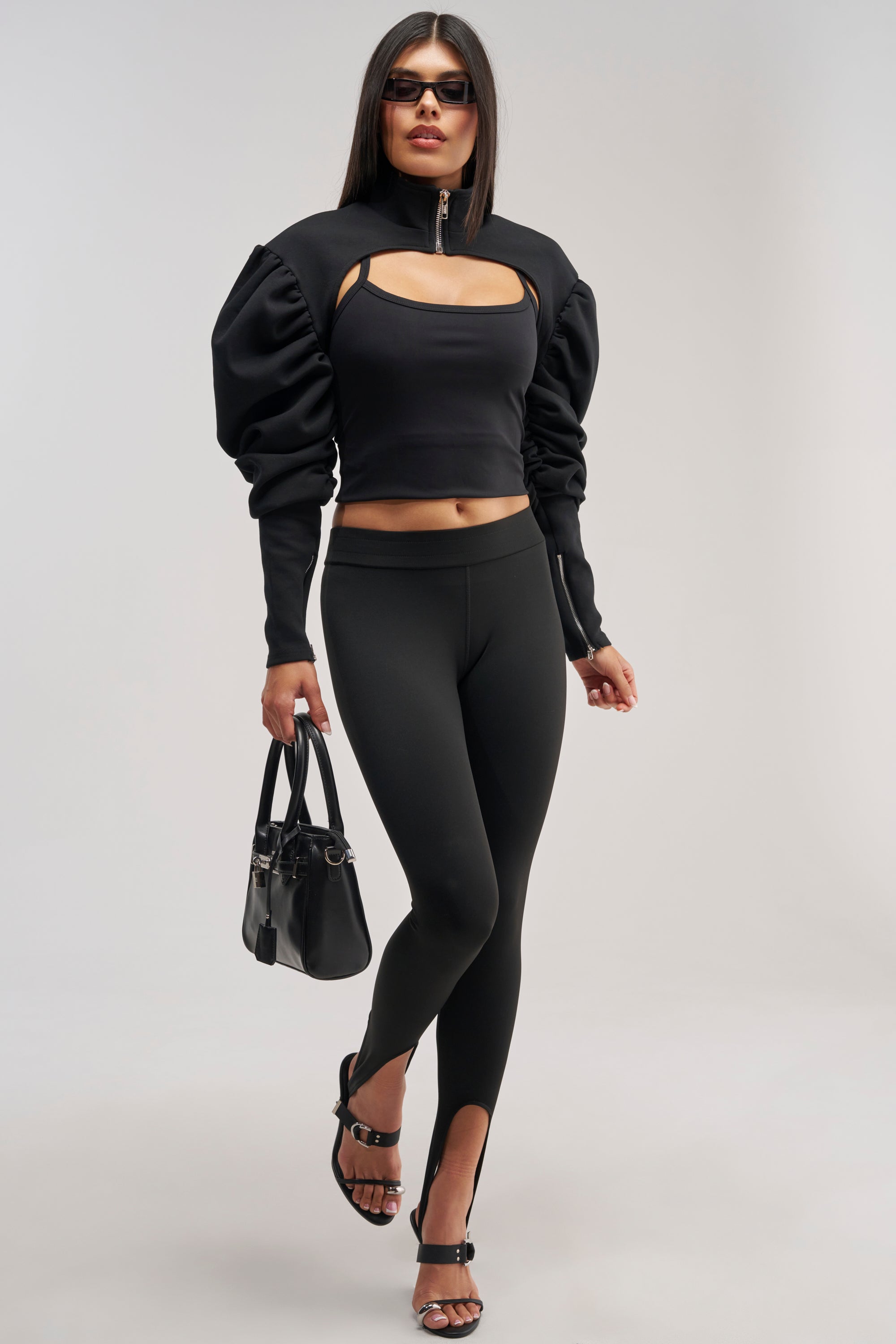 A woman strides confidently against a light gray background, wearing a black cut-out top with puff sleeves, heeled sandals, sunglasses, a black handbag, and THE FEMME FATALE STIRRUP LEGGING.