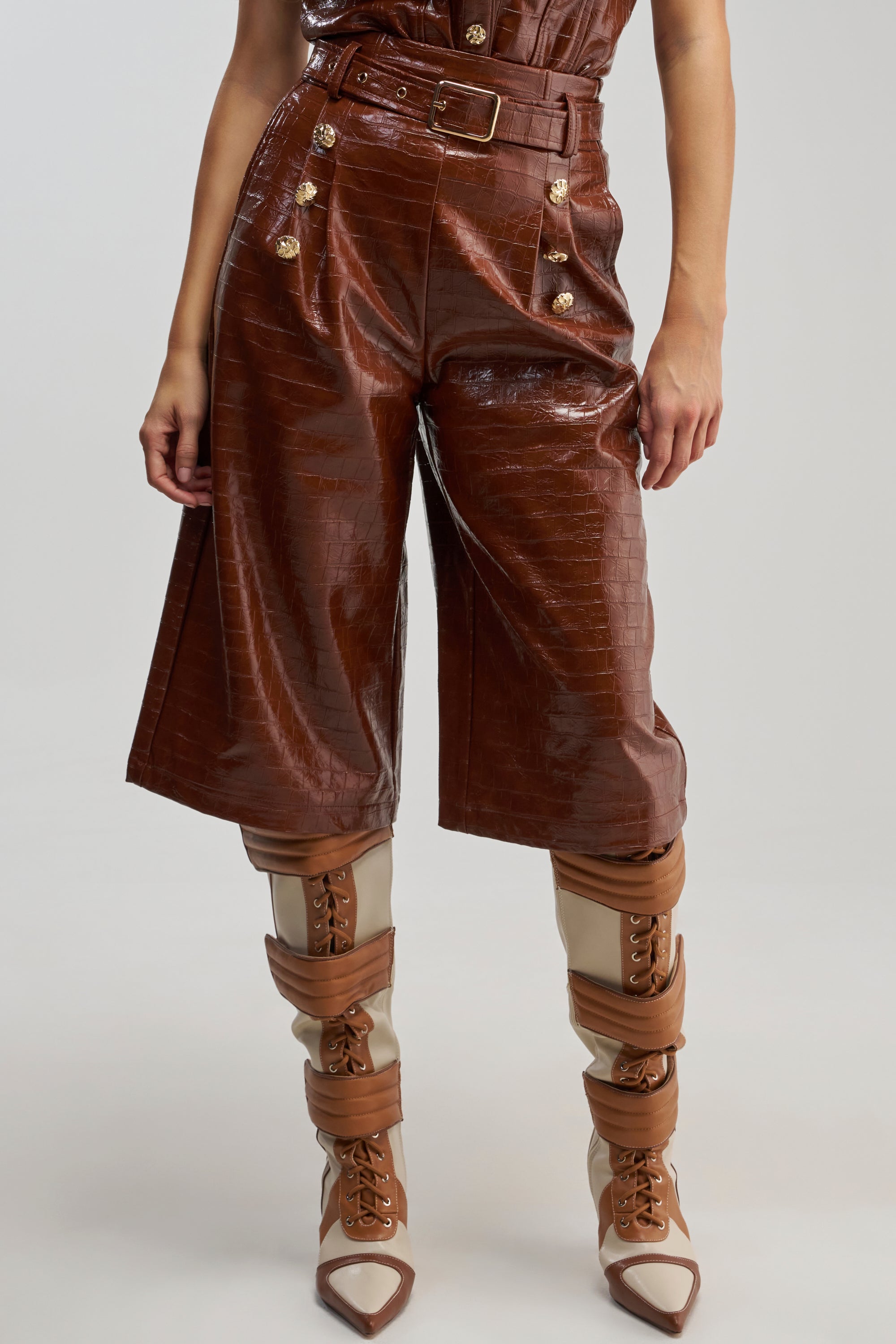 A person is wearing the WOMAN IN POWER TROUSER, featuring shiny brown high-waisted faux leather culotte pants with gold buttons and a belt, paired with tan and brown lace-up heeled boots.