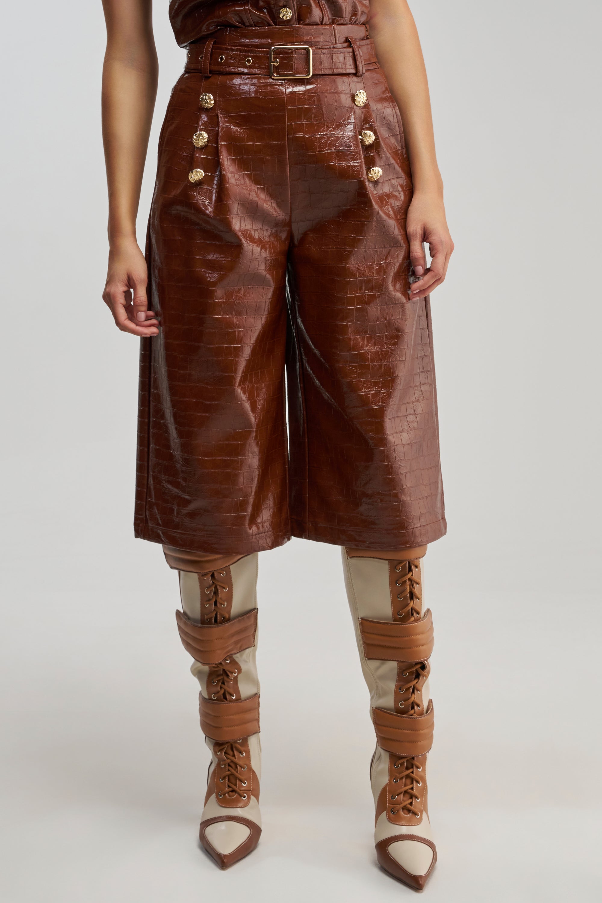 A person wearing the WOMAN IN POWER TROUSER—high-waisted, brown, crocodile-embossed culottes with gold buttons—paired with beige and brown high-heeled boots for an elevated take on officewear.