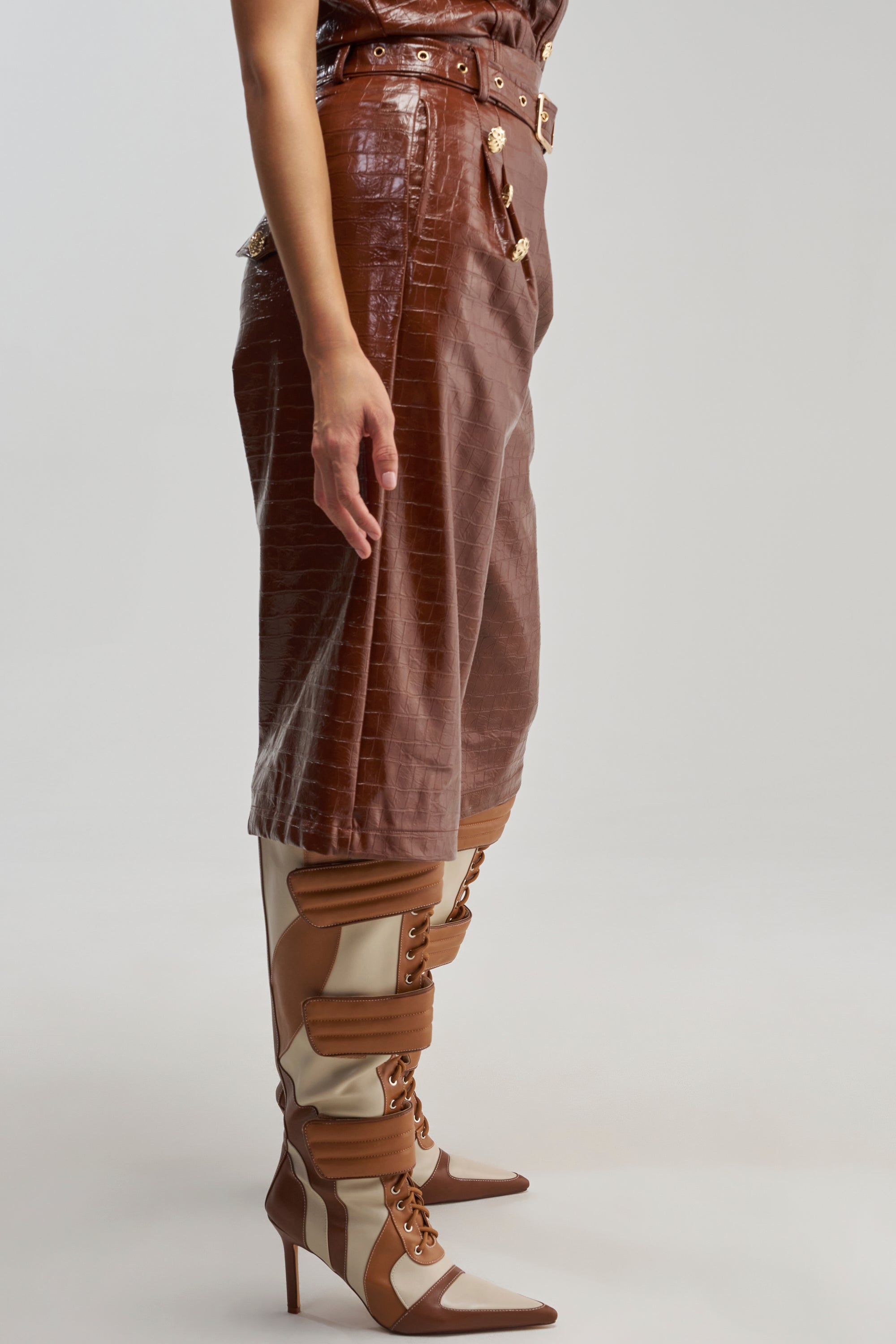 Clad in the WOMAN IN POWER TROUSER—brown faux leather with a crocodile-embossed finish—and tan-and-cream high-heeled lace-up boots, this figure stands against a plain gray background. The image focuses on the lower half of the body, showcasing a modern approach to officewear style.