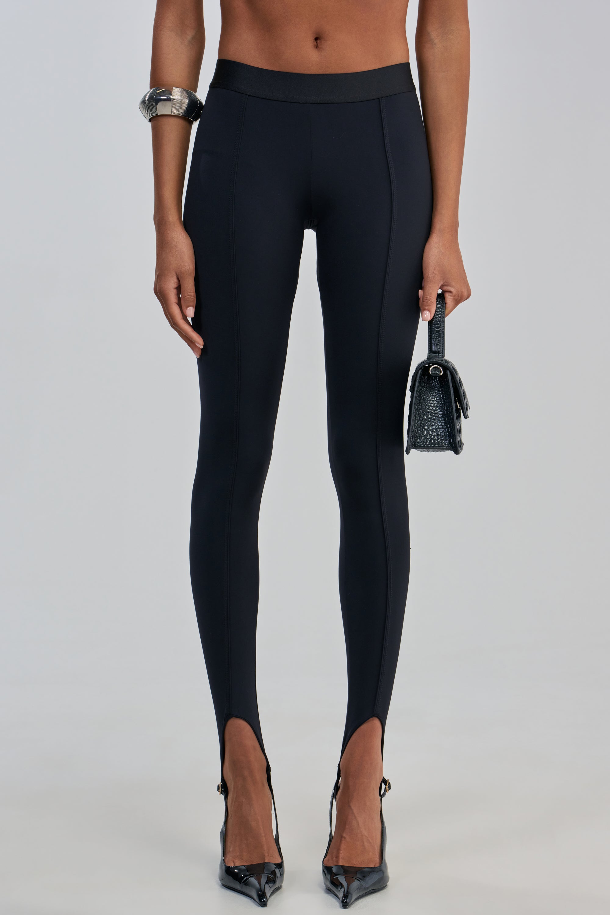 A person wearing THE BEST STIRRUP LEGGING styled with black pointed high heels, a chunky bracelet, and holding a small textured handbag against a plain background.