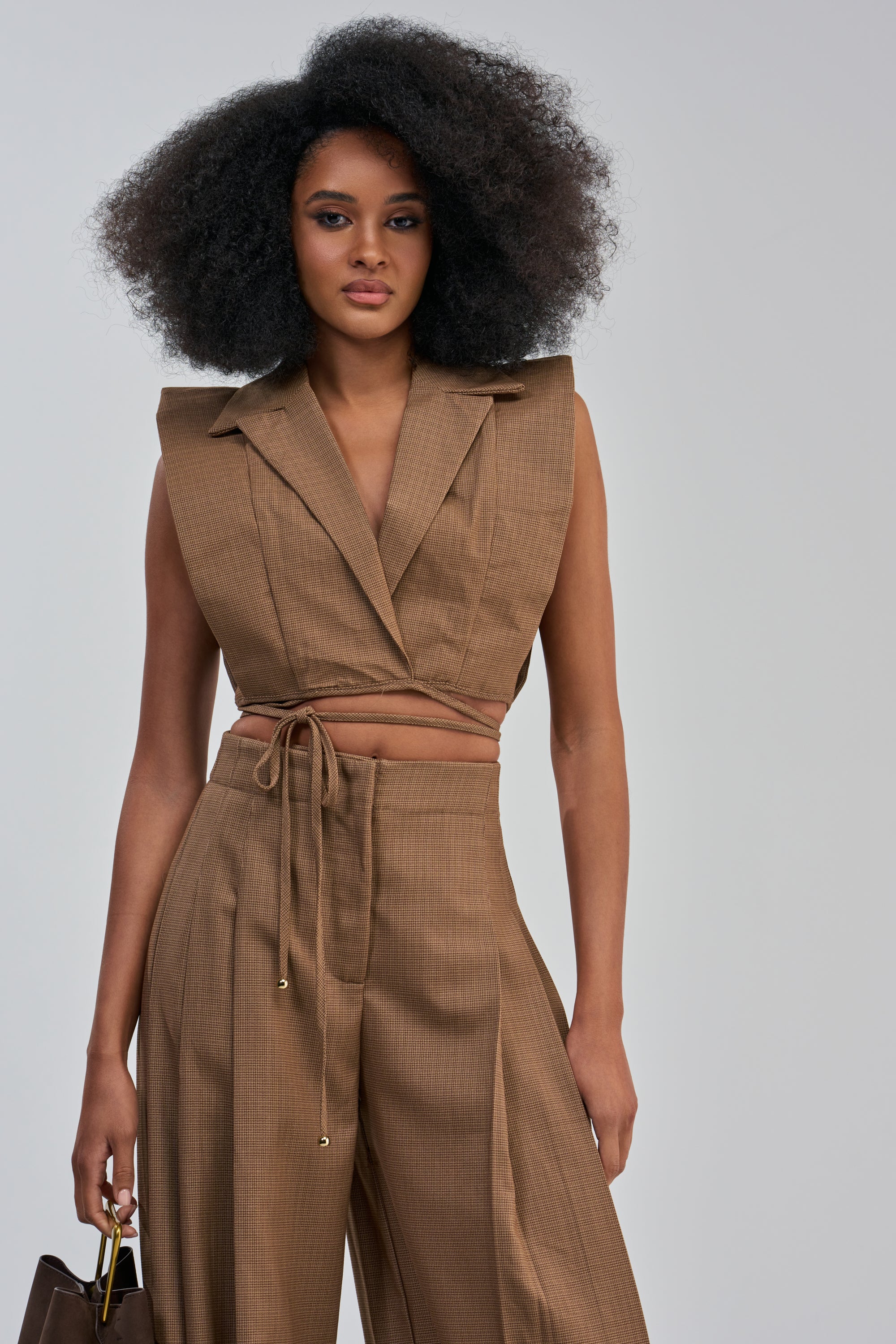 A woman stands facing forward in the GUESS WHO V-NECK COLLARED BLOUSE IN BROWN, paired with wide-leg pants and a matching brown handbag—a striking example of modern power-dressing. Her voluminous afro and the plain light gray backdrop complete the bold, contemporary look.