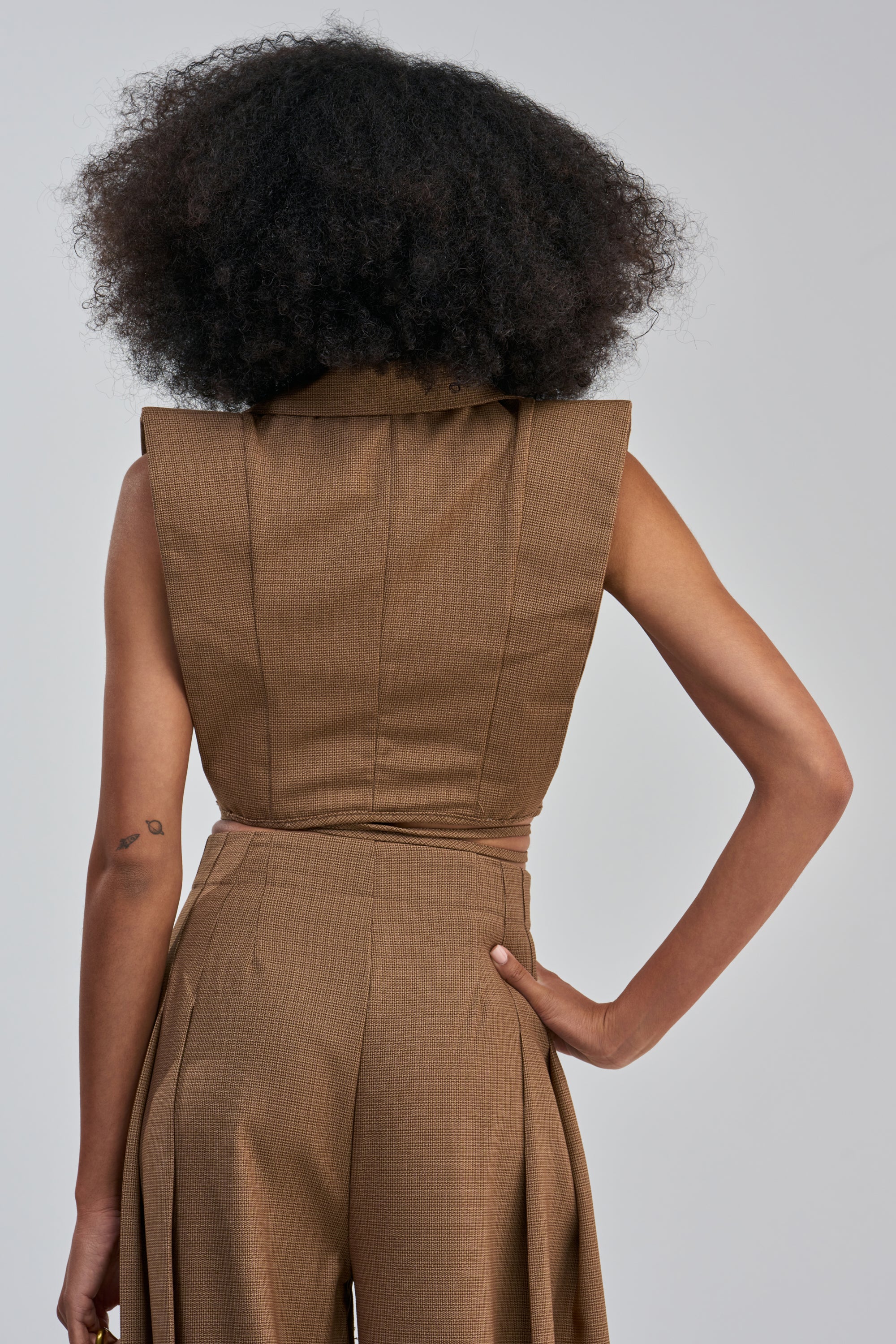 A person with curly hair stands facing away, channeling power-dressing in the GUESS WHO V-NECK COLLARED BLOUSE IN BROWN and matching high-waisted pants. One hand rests on their hip, revealing a small tattoo on the upper arm, while the plain gray background highlights this modern approach to workwear.
