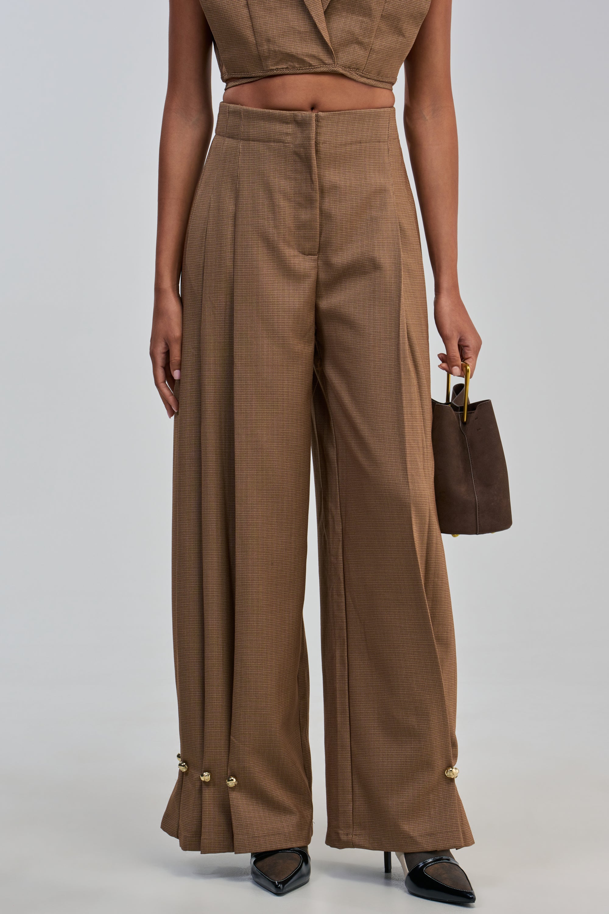 Wearing the GUESS WHO BALLOON PANT IN BROWN—a high-waisted, wide-leg silhouette featuring gold buttons at the hem—paired with a matching cropped top, black pointed shoes, and a small brown handbag creates an effortlessly office-ready look.