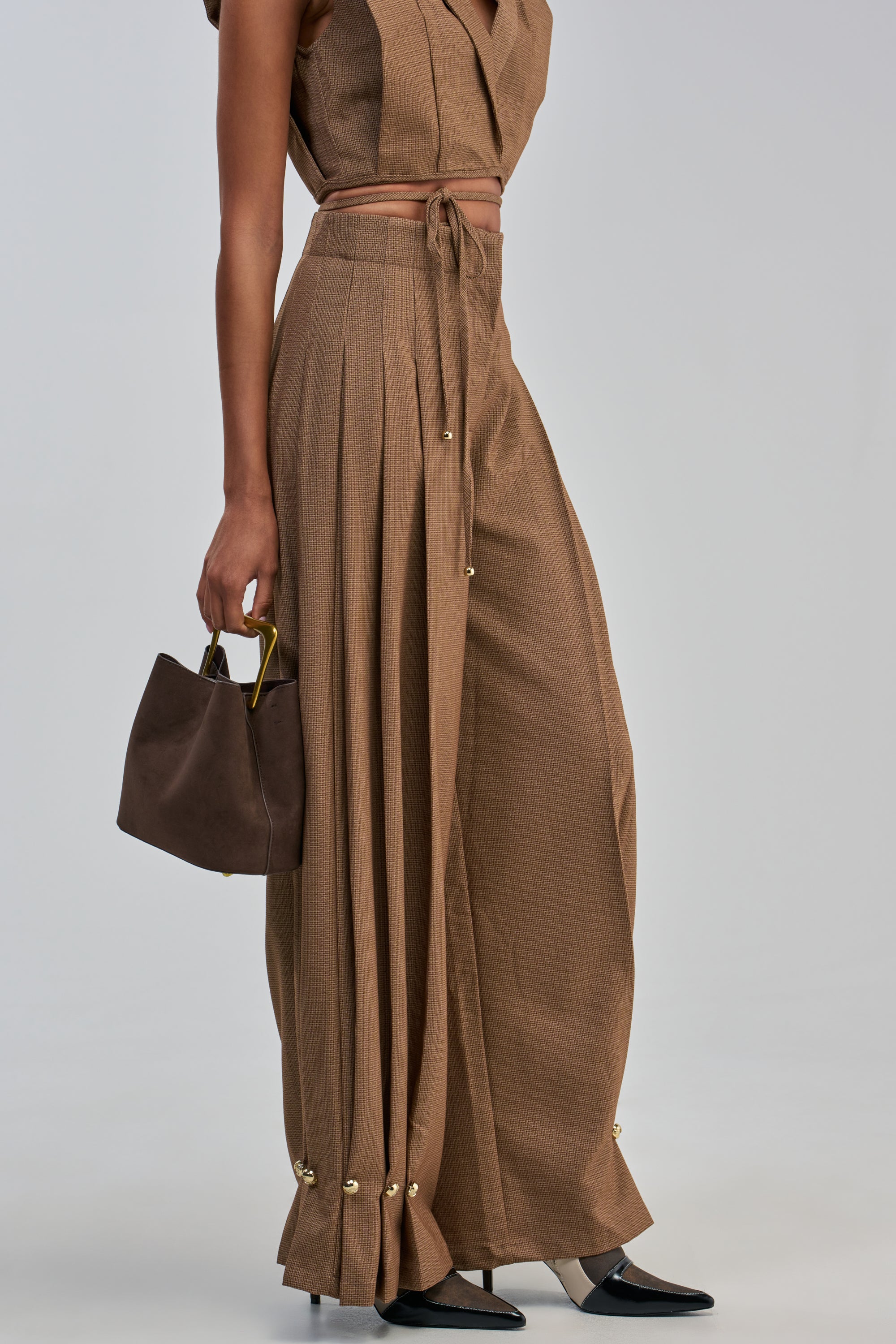Wearing the GUESS WHO TROUSER—a wide-leg silhouette in brown—paired with a matching cropped top, gold accents, gray and black heeled shoes, and carrying a small dark brown handbag, creates an effortlessly office-ready look.