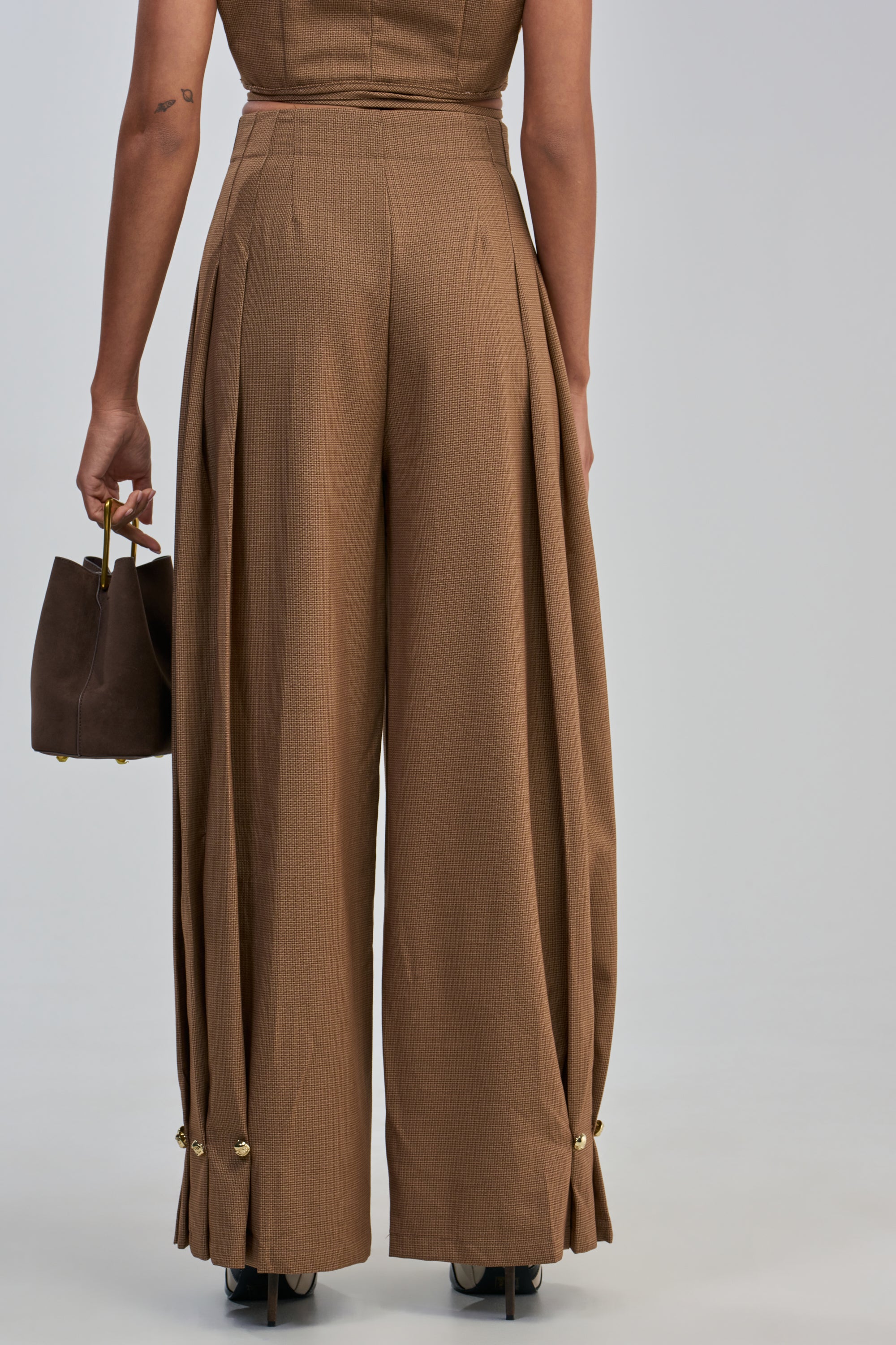 A person stands facing away, wearing the GUESS WHO TROUSER—office-ready, high-waisted, wide-leg brown trousers featuring pleats and gold buttons at the ankles—and holds a brown handbag in their left hand.