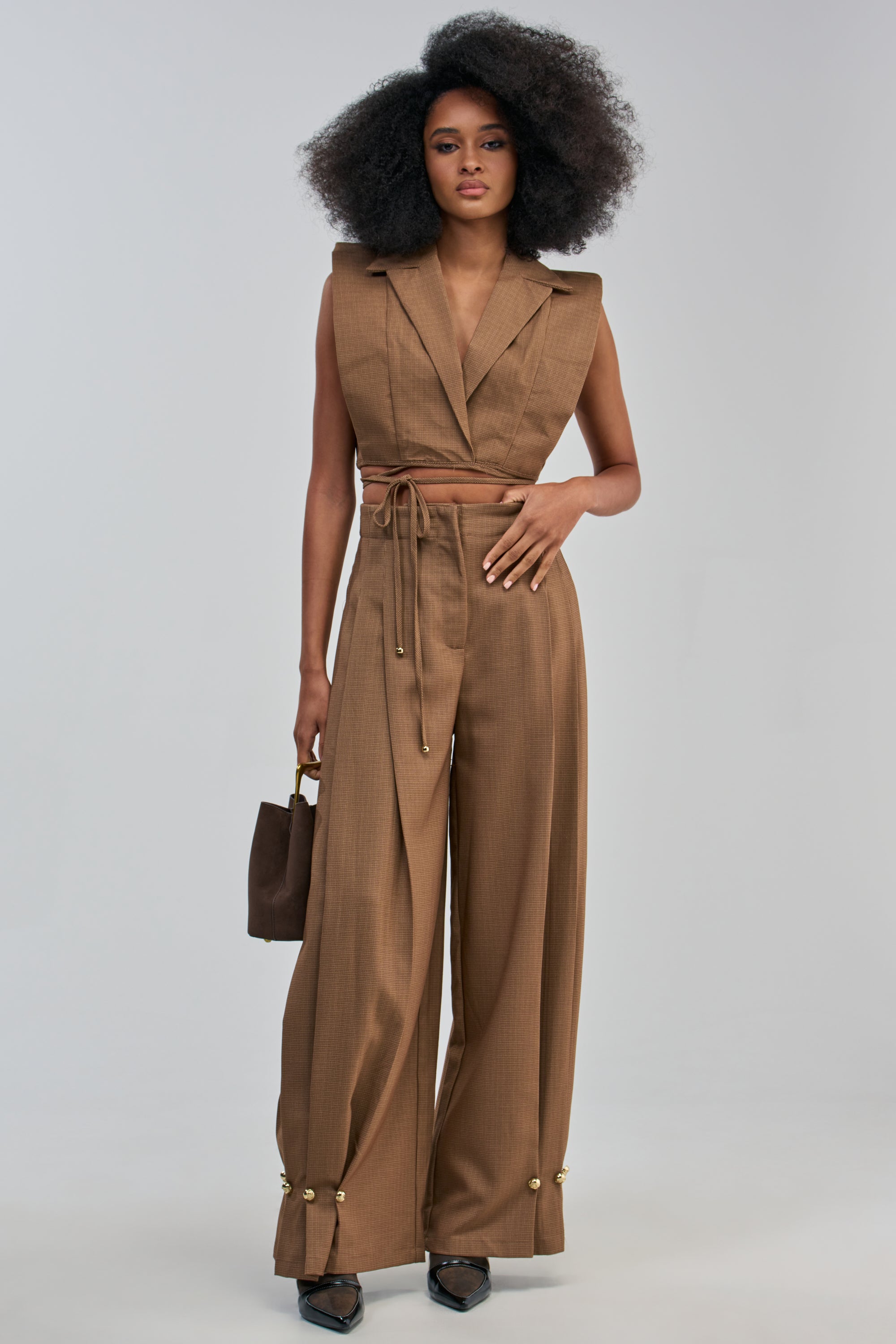 A person wearing the GUESS WHO TROUSER, an office-ready sleeveless brown suit set featuring wide-leg pants with a belted waist and gold accents at the ankles, holds a small brown purse. The elegant ensemble is set against a plain, light background.