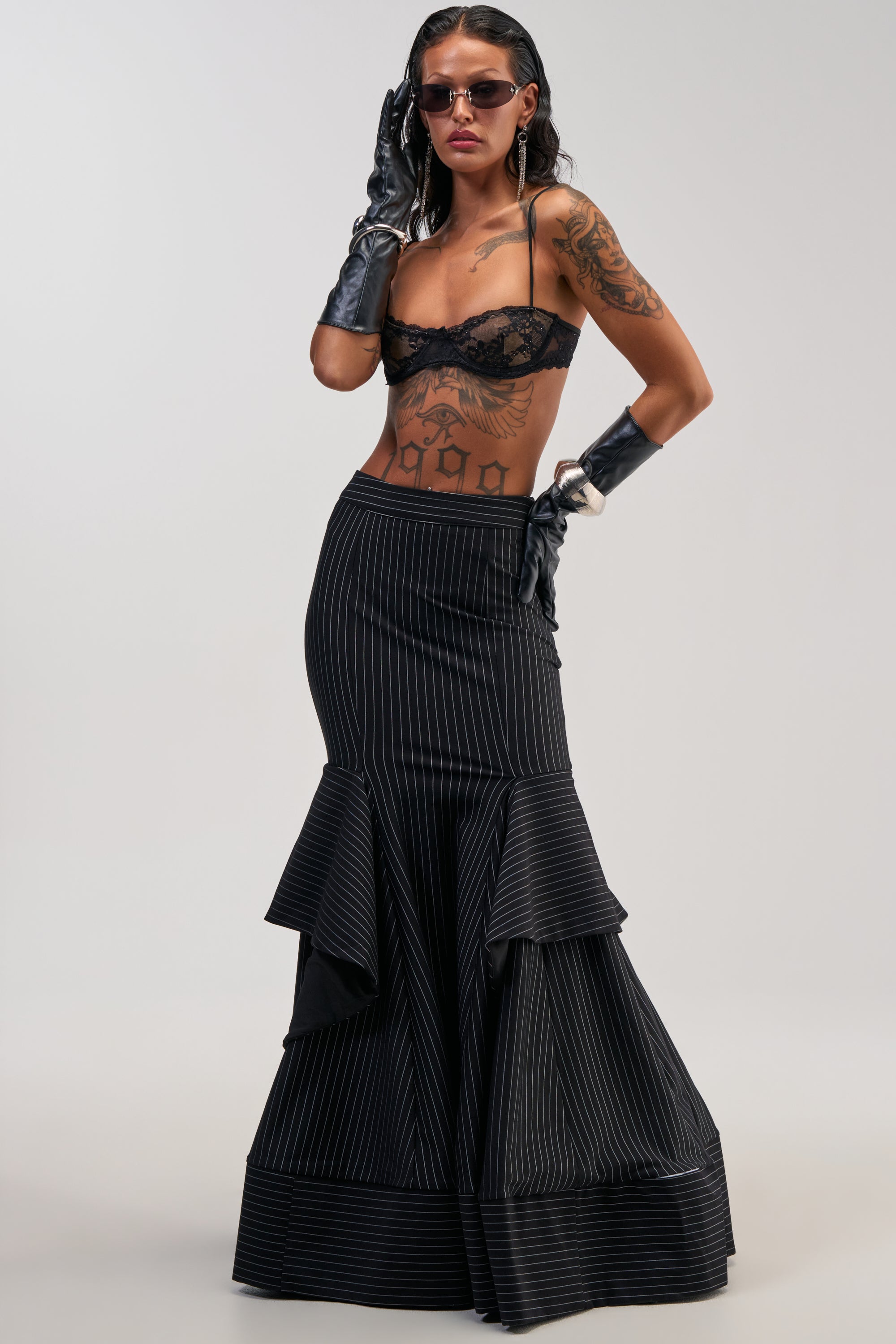 A woman with tattoos poses in sunglasses, a black lace bralette, and the GIVE IT UP PINSTRIPE MAXI SKIRT with a ruffle hem, finished with black gloves—capturing the bold confidence of the office siren aesthetic against a plain background.
