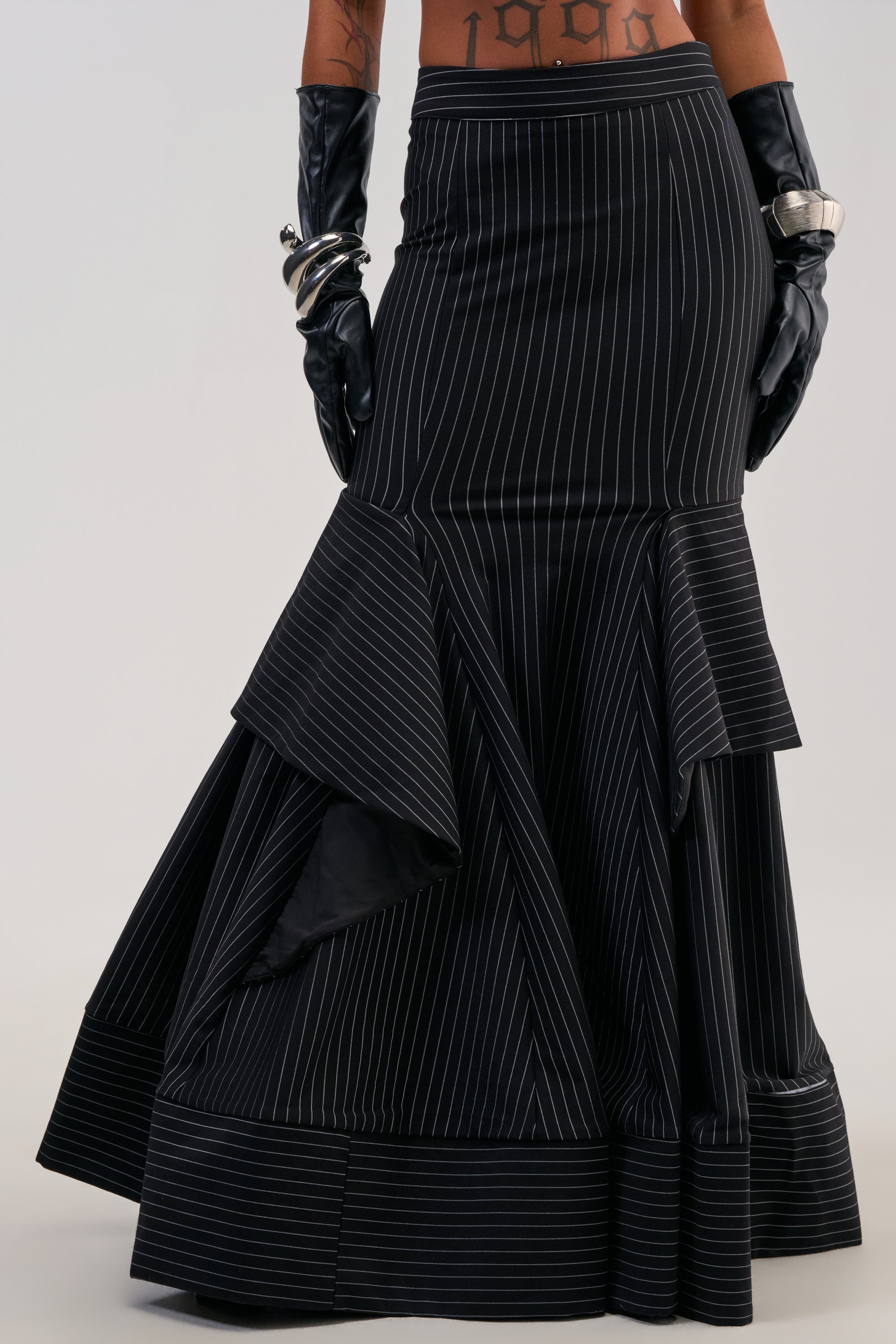Wearing the GIVE IT UP PINSTRIPE MAXI SKIRT—an office siren-inspired black pinstripe maxi skirt featuring dramatic flares and a ruffle hem—styled with black leather gloves, silver bangles, and visible tattoos above the waistline.