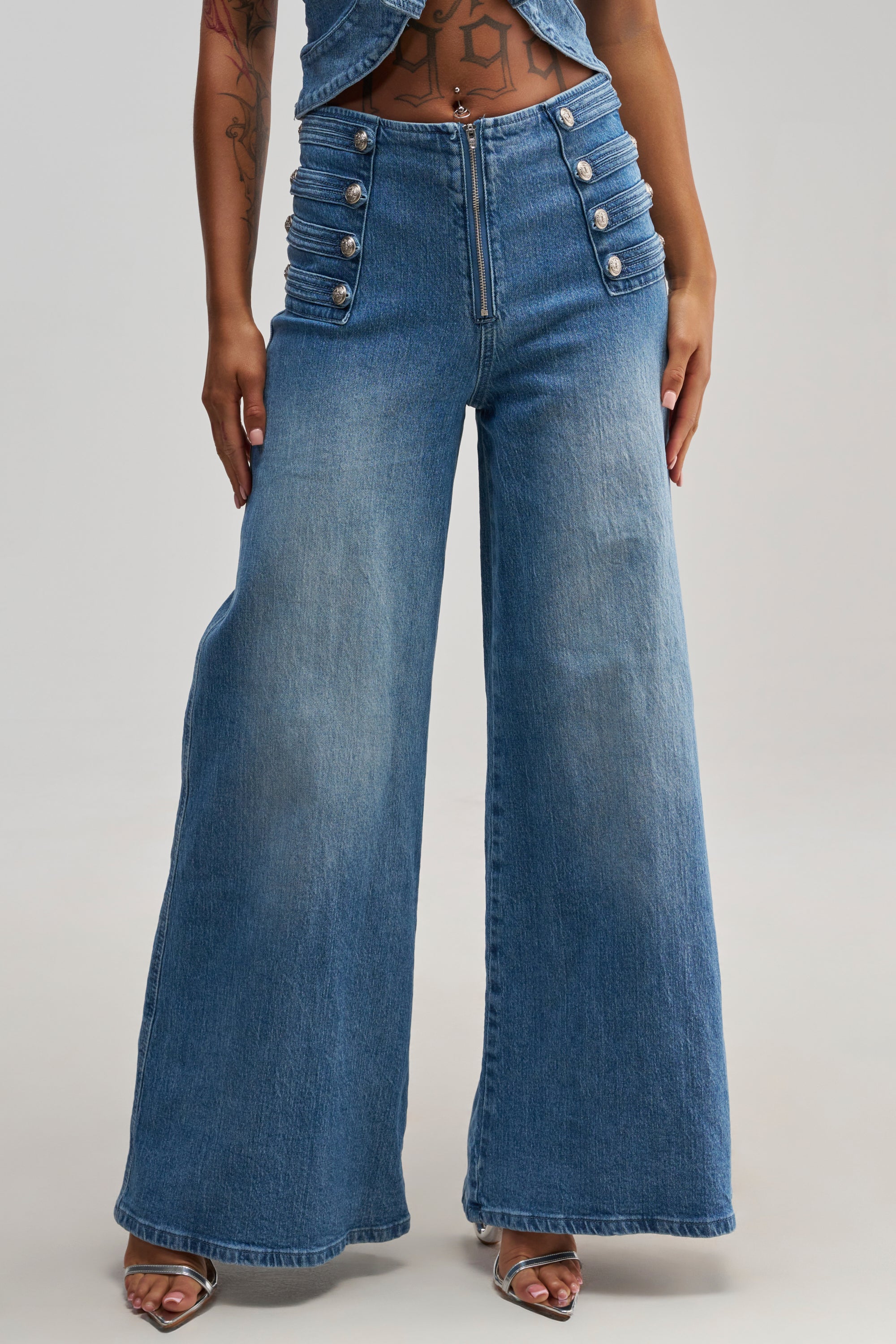 A person wearing the ALIGNED MILITARY DENIM PANT—high-waisted, wide-leg jeans featuring multiple decorative buttons on each hip—stands in open-toe heels against a plain background.