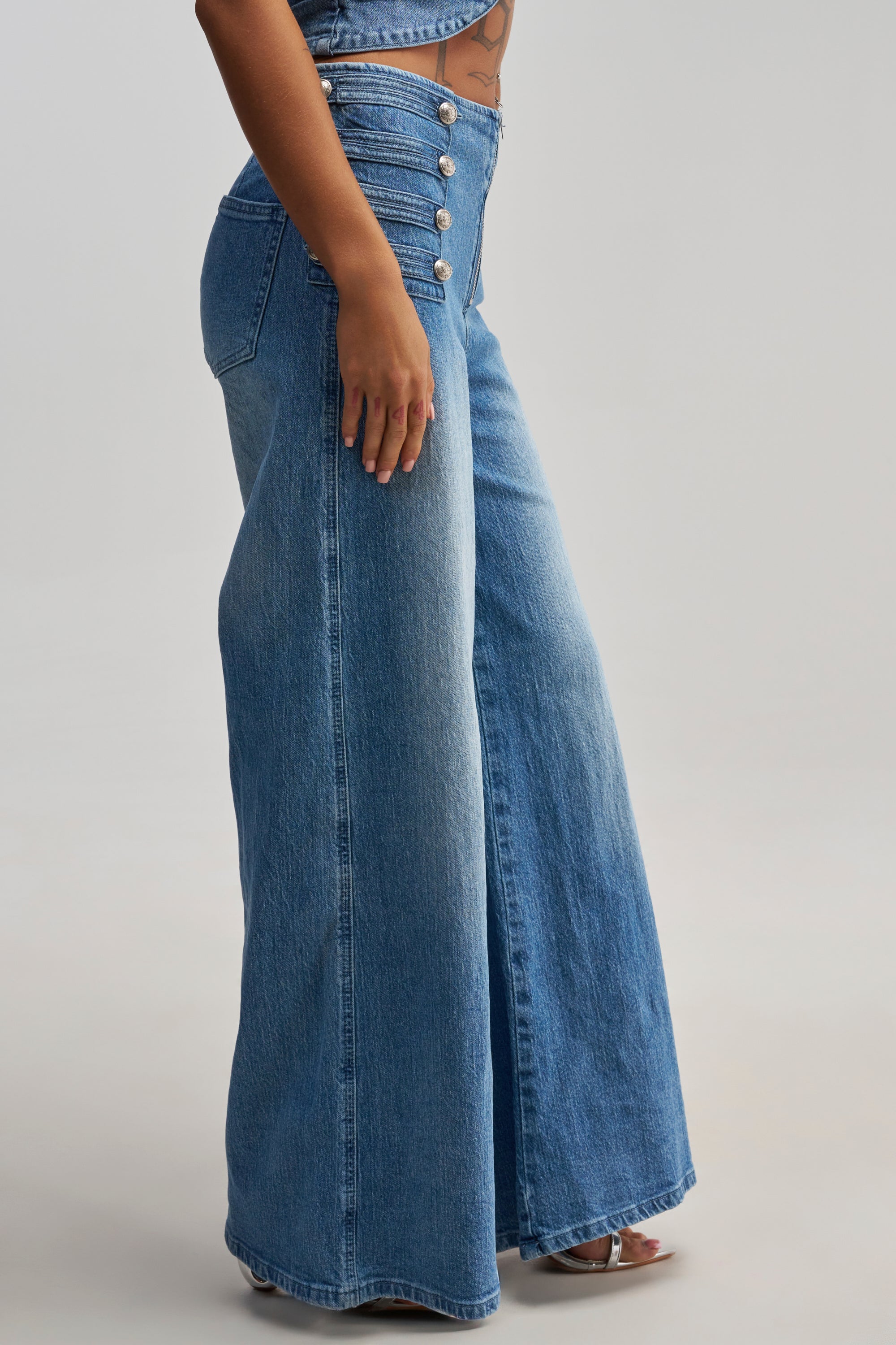 Wearing the ALIGNED MILITARY DENIM PANT—a high-waisted, wide-leg blue jean accented with multiple silver buttons on the front—a person stands sideways against a plain background, embodying a core streetwear style.