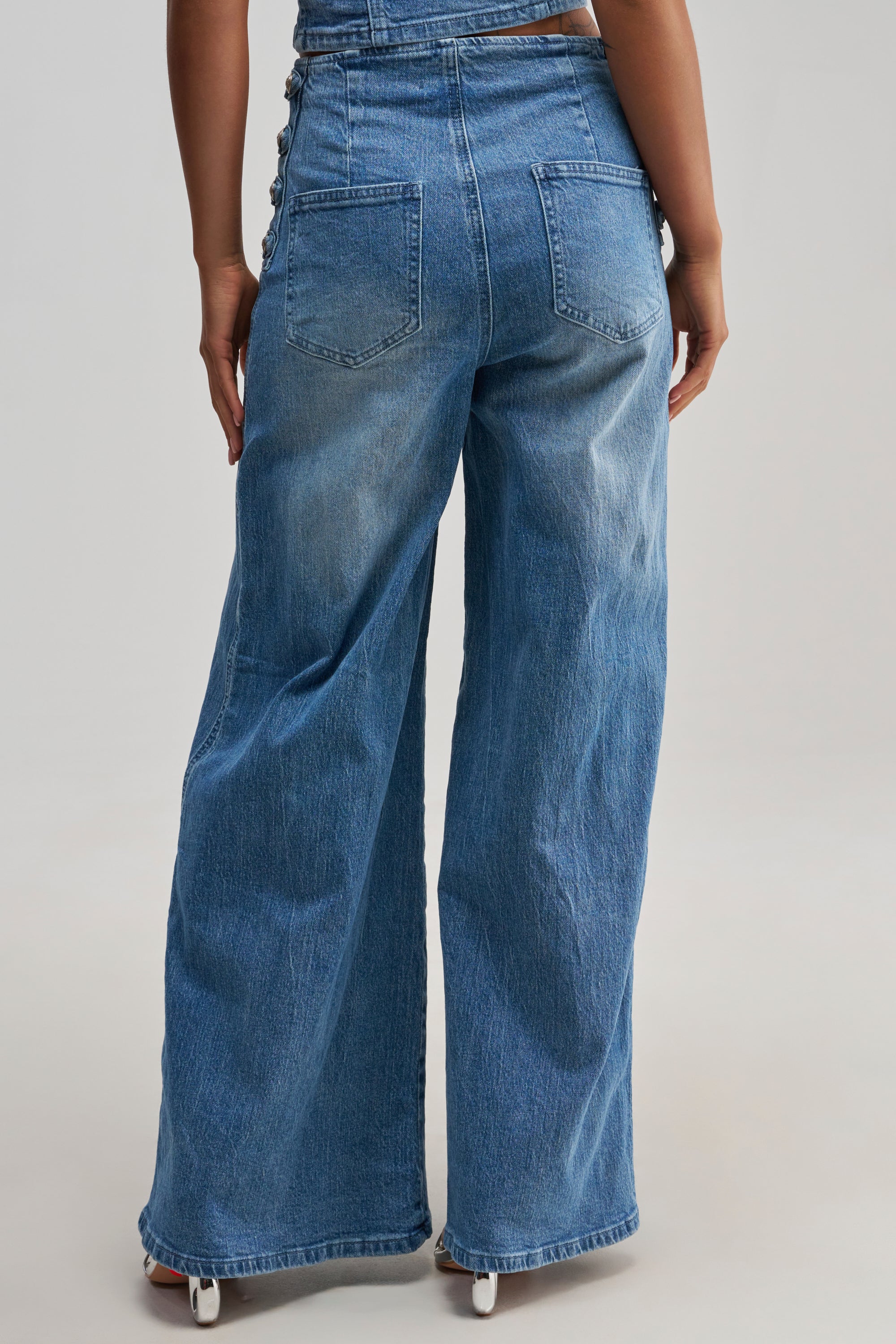 A person is shown from behind against a plain light background, wearing the ALIGNED MILITARY DENIM PANT. These high-waisted, wide-leg jeans feature back pockets, visible seam details, and a relaxed fit that channels an effortless streetwear vibe.