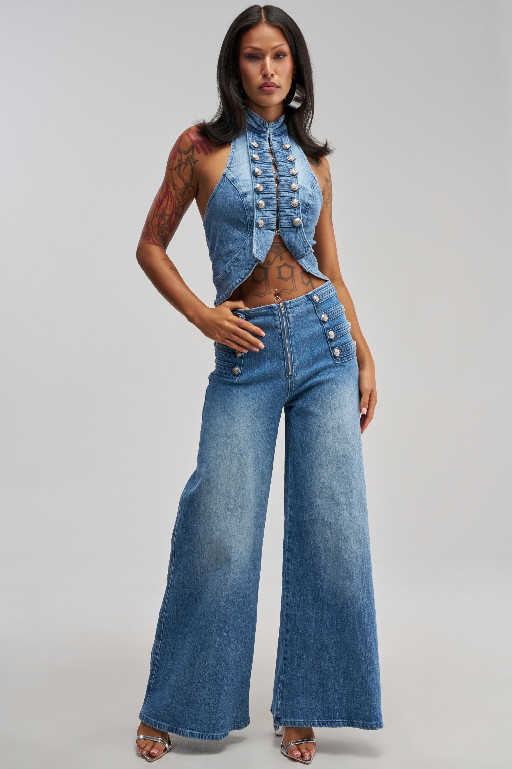Wearing the ALIGNED MILITARY DENIM PANT, the woman pairs these high-waisted, wide-leg jeans with a sleeveless, high-neck denim crop top adorned with decorative buttons. She stands confidently with one hand on her hip and the other resting by her waist.
