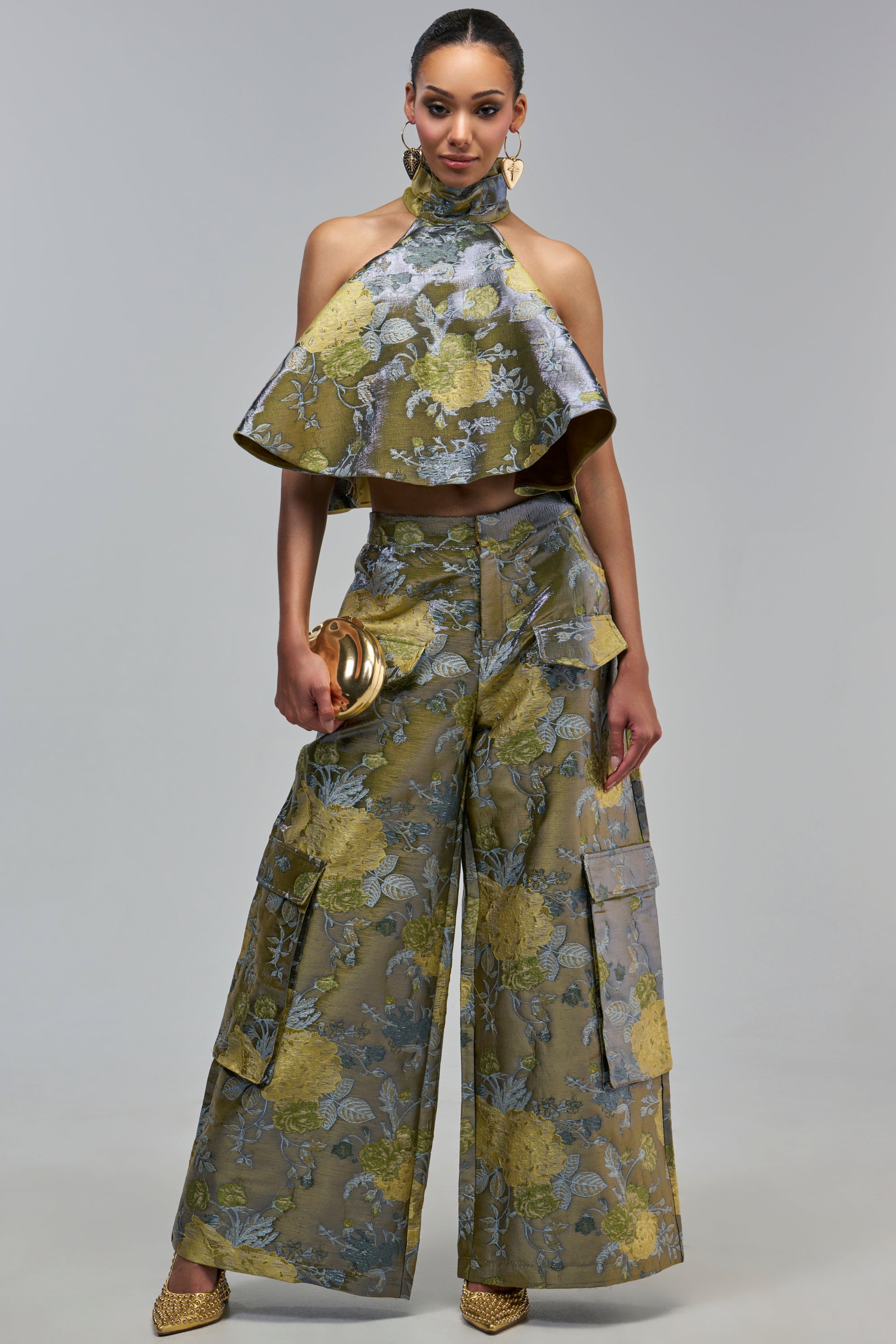 A model wears the BEAUTIFUL IN BROCADE TROUSER set in olive green floral brocade, featuring a halter-neck crop top and wide-leg trousers, accessorized with a gold clutch and heart-shaped earrings for statement fall styling.