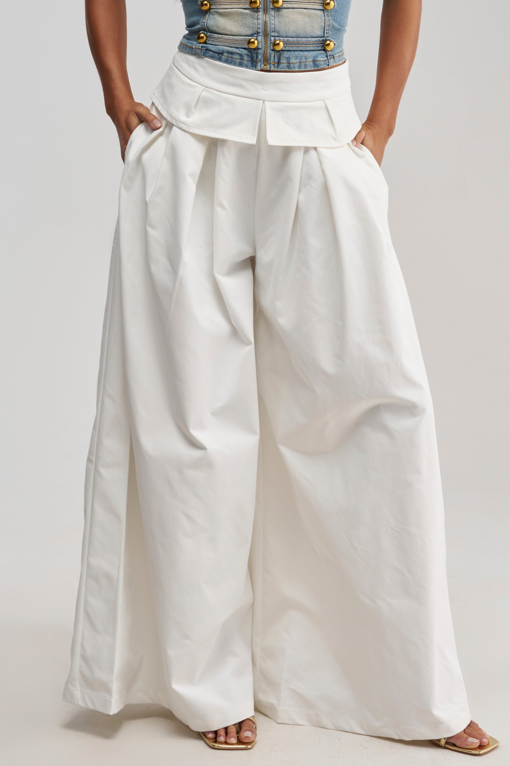 A person wearing the LOCKED IN LOVE TROUSER—high-waisted, wide-leg white pleated trousers that are flowy and elegantly tailored—paired with a denim top featuring gold buttons and open-toed heels, stands with hands in their pockets.