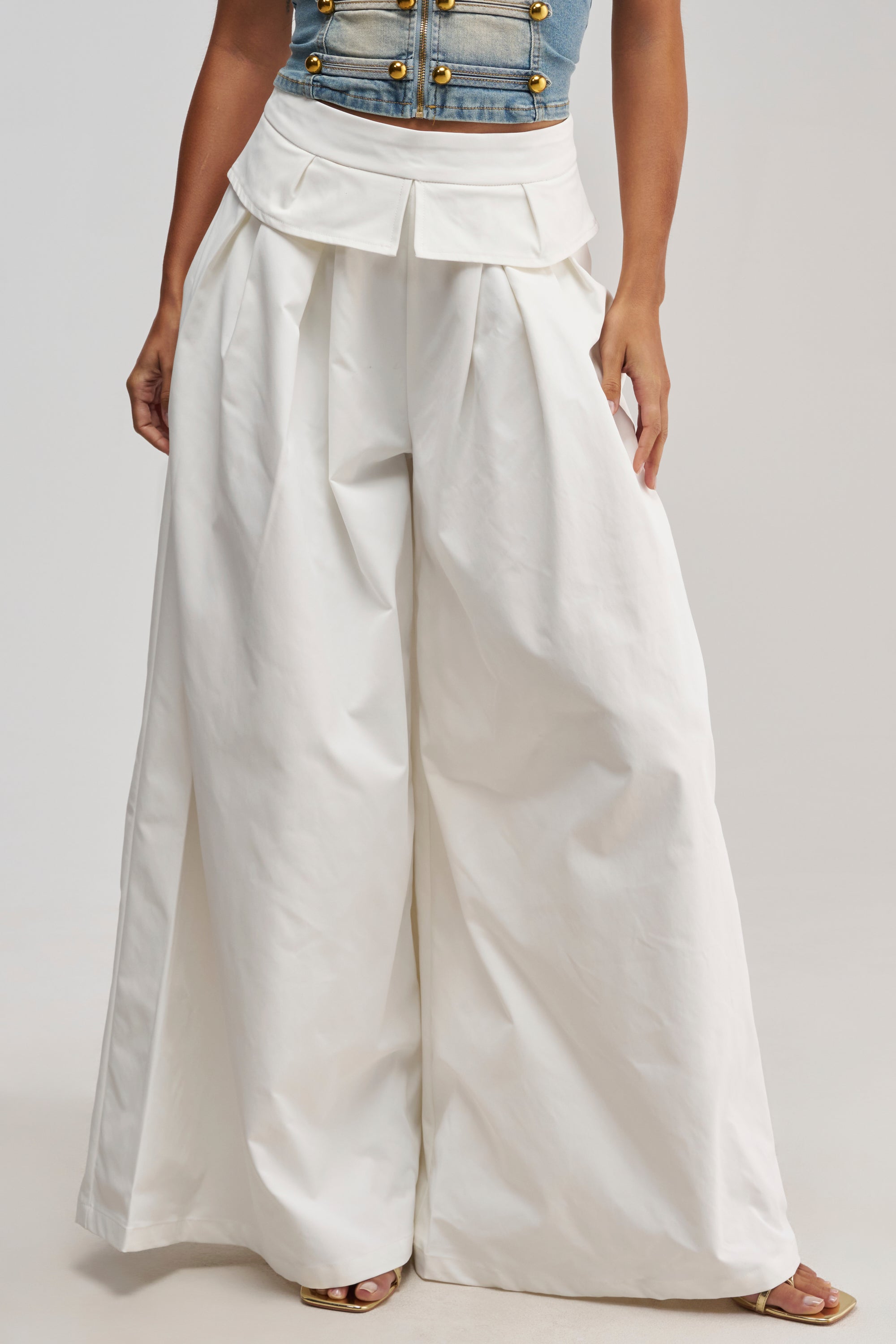 Wearing the LOCKED IN LOVE TROUSER, a pair of high-waisted, wide-leg white trousers featuring pleats and a distinctive structured peplum waist, the person pairs them with a denim top adorned with gold buttons and stands with hands in their pockets.