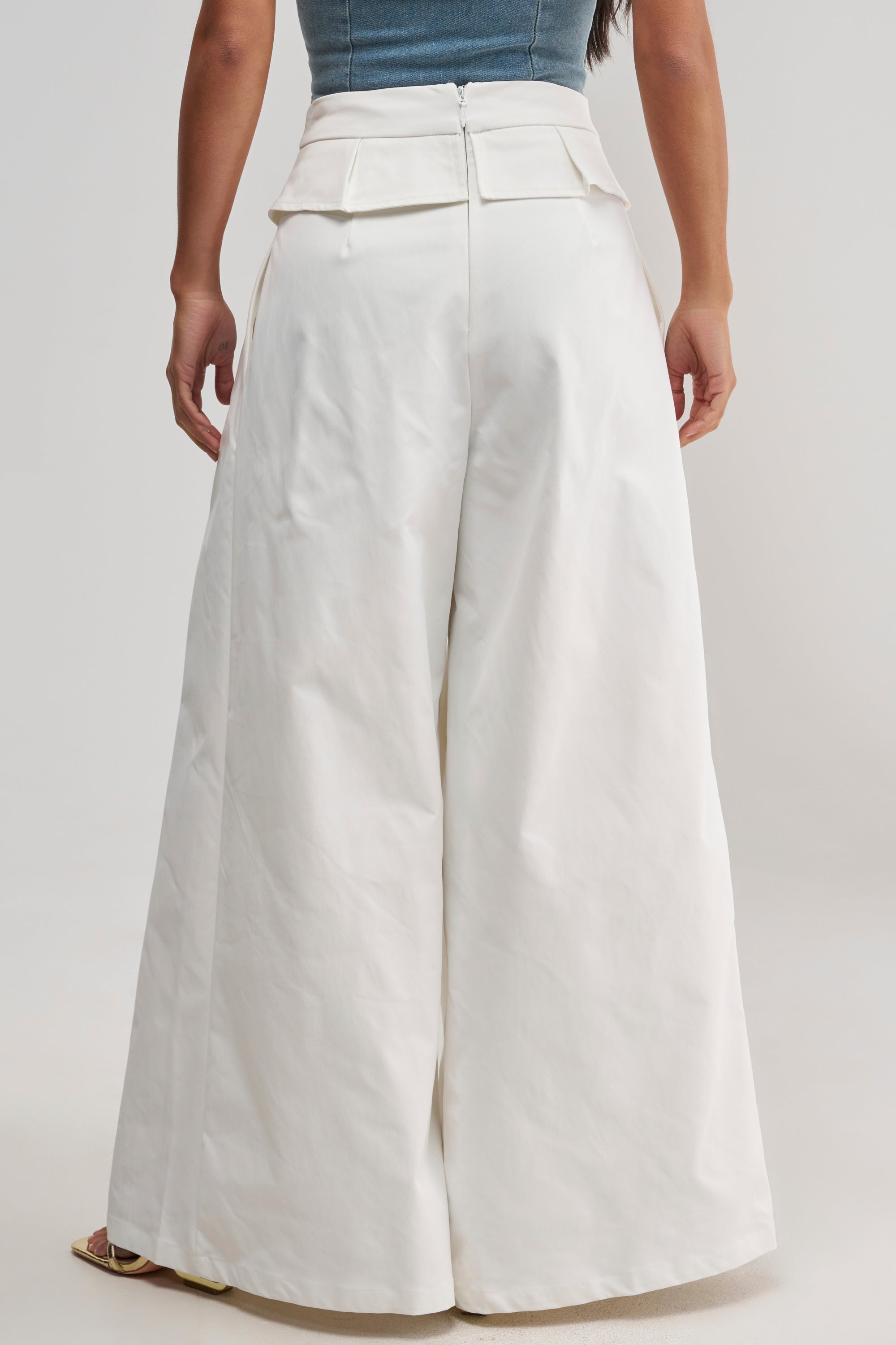 A person wearing the LOCKED IN LOVE TROUSER—high-waisted, wide-leg white trousers with a zipper and flap detail on the back—paired with a tucked-in blue top, is standing facing away from the camera.