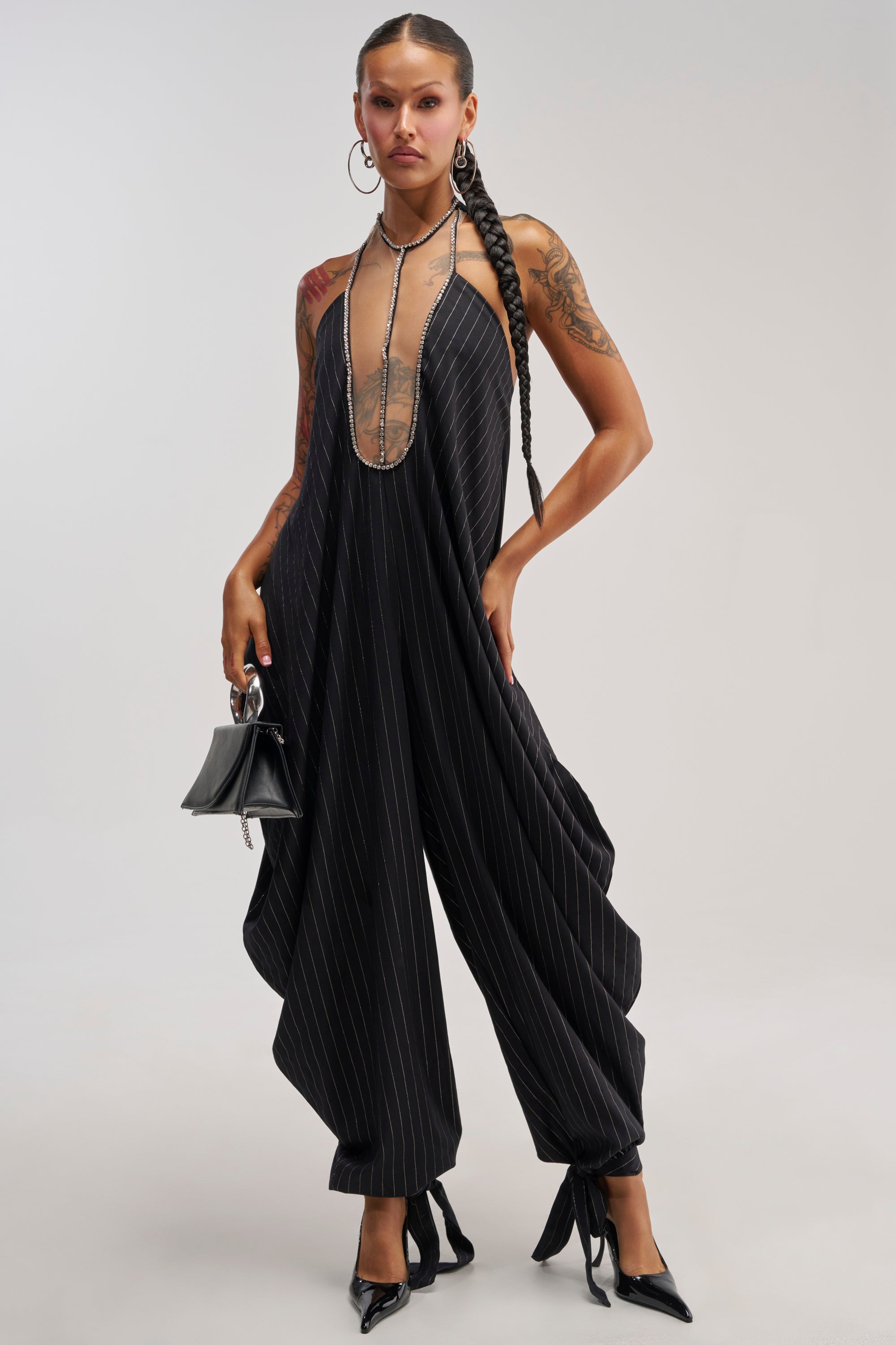 Wearing the READY FOR BUSINESS PINSTRIPE JUMPSUIT, this person stands against a plain background. The loose, black pinstripe jumpsuit features a deep neckline. They accessorize with a small black handbag, hoop earrings, and pointed black heels. Their look is completed with long braids and visible tattoos.