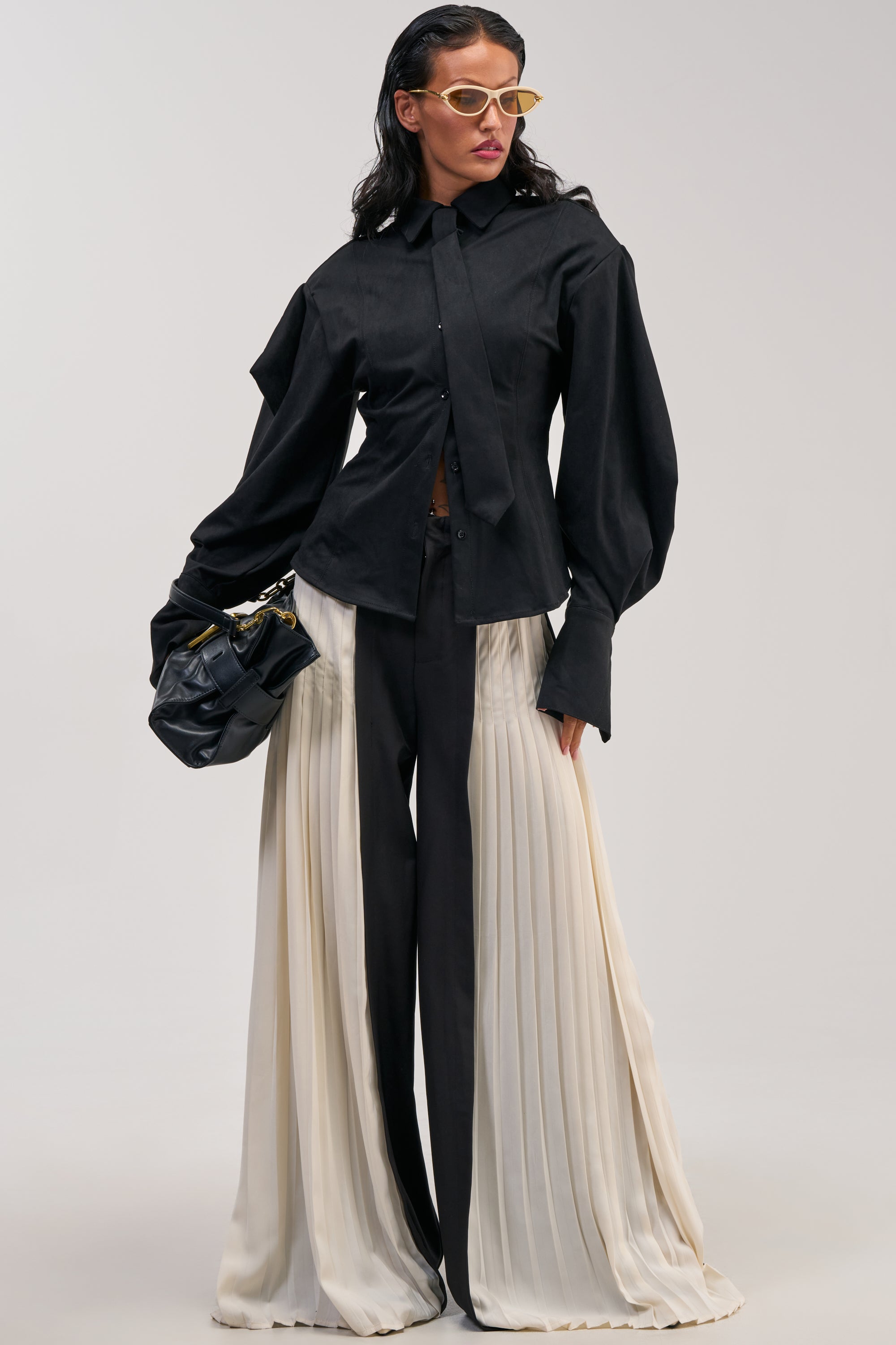 A person poses in a black button-up shirt and the MIXED VIBES TROUSER—black pleated panel trousers featuring wide cream fabric panels and a high-rise waist—styled with yellow-tinted glasses and a black clutch for an effortlessly chic look, perfect for cocktail parties. The background is plain and light-colored.