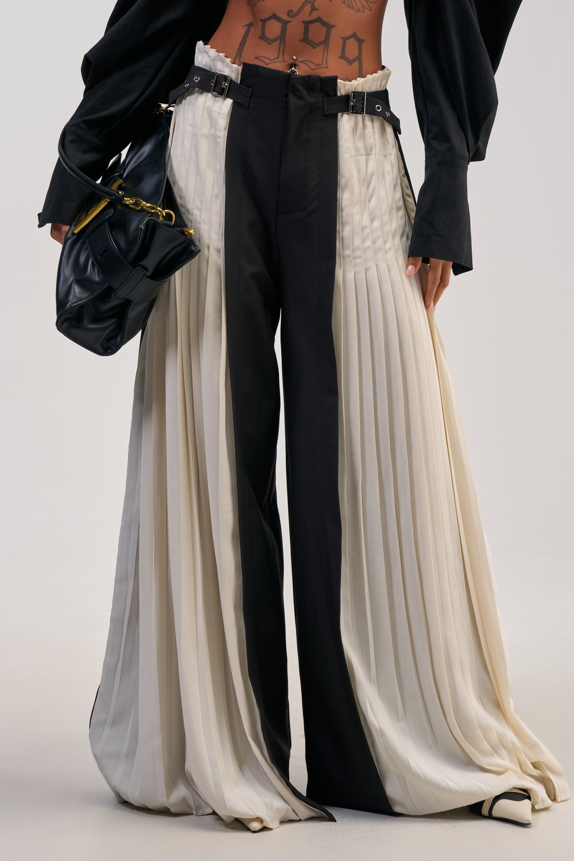 Wearing the MIXED VIBES TROUSER—high-waisted black and cream pleated panel trousers with a striking high-rise waist—a person showcases their “1999” lower torso tattoo while carrying a black handbag, making it the perfect choice for stylish cocktail parties.