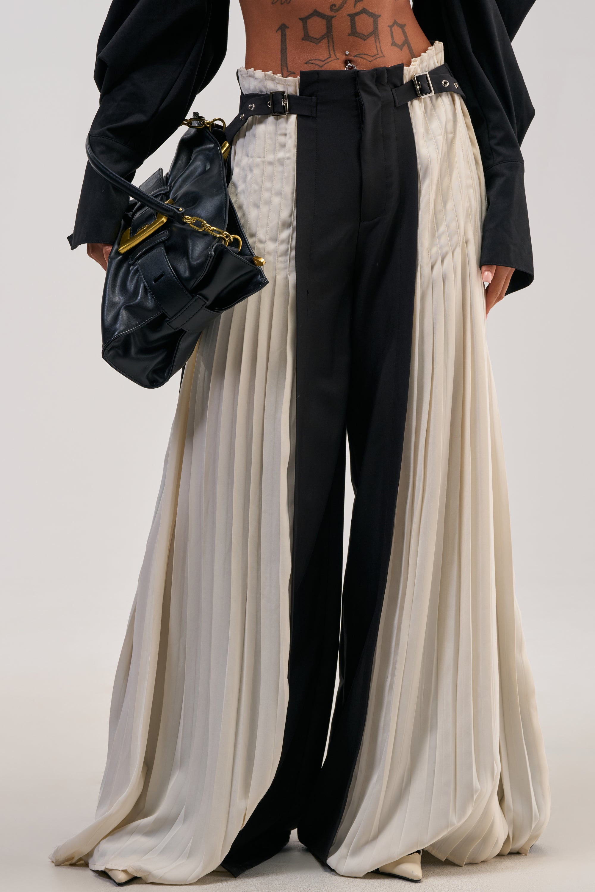 Wearing the MIXED VIBES TROUSER—featuring black and cream pleated panels with a high-rise waist—paired with a black jacket and accessorized with a black handbag. A “1999” torso tattoo is visible, adding an edge to this stylish, fashion-forward look that’s perfect for cocktail parties. The ensemble stands out against a plain background.