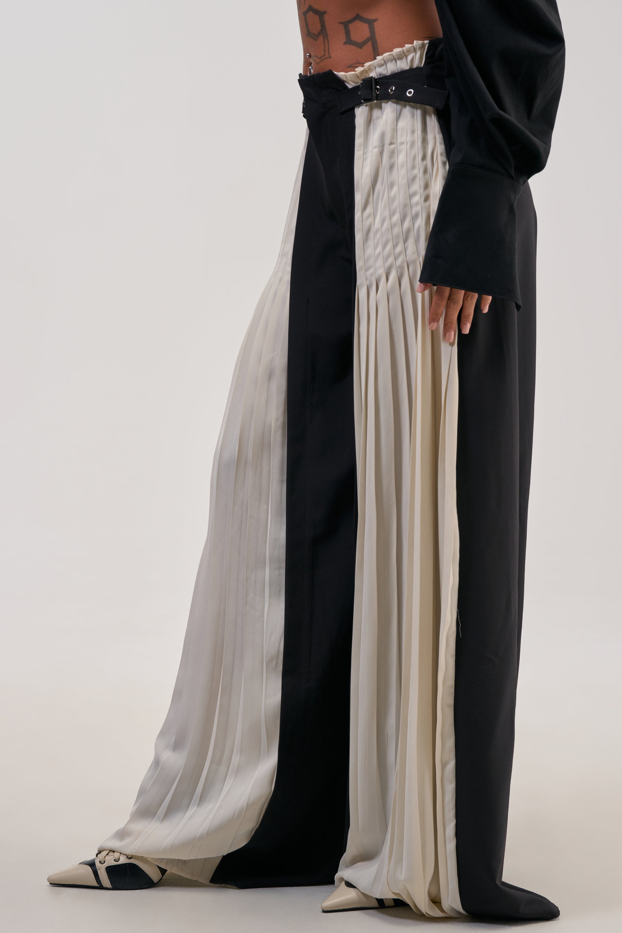 Wearing the MIXED VIBES TROUSER—featuring black and white pleated panels, a high-rise belted waist—and paired with pointed shoes and a visible torso tattoo, this look is perfect for making a statement at cocktail parties.