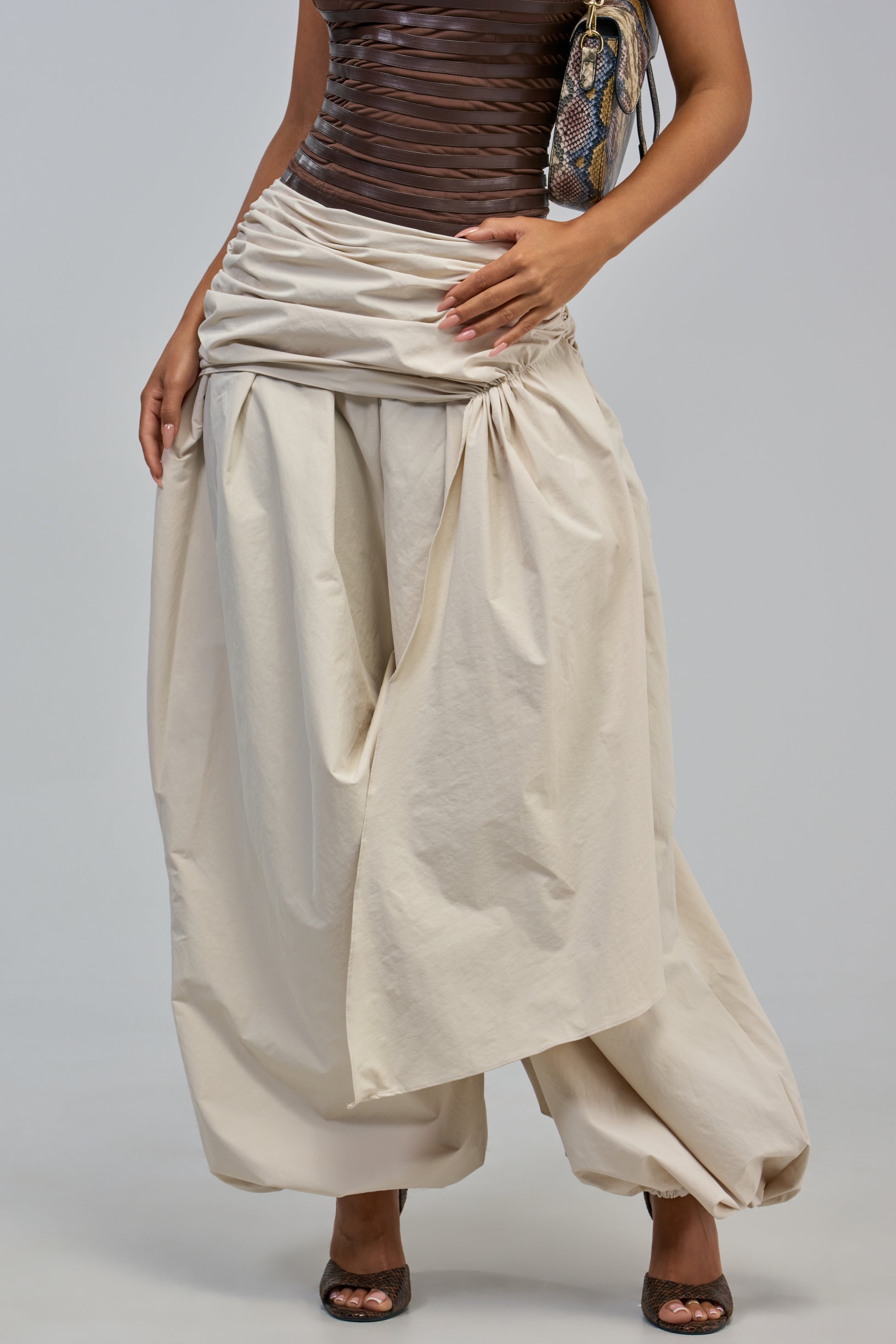 A person wearing the NOT MY TYPE TROUSER—loose, draped balloon trousers in beige fabric—paired with a fitted brown top and carrying a snakeskin-patterned bag. Only the lower half of their body is visible.