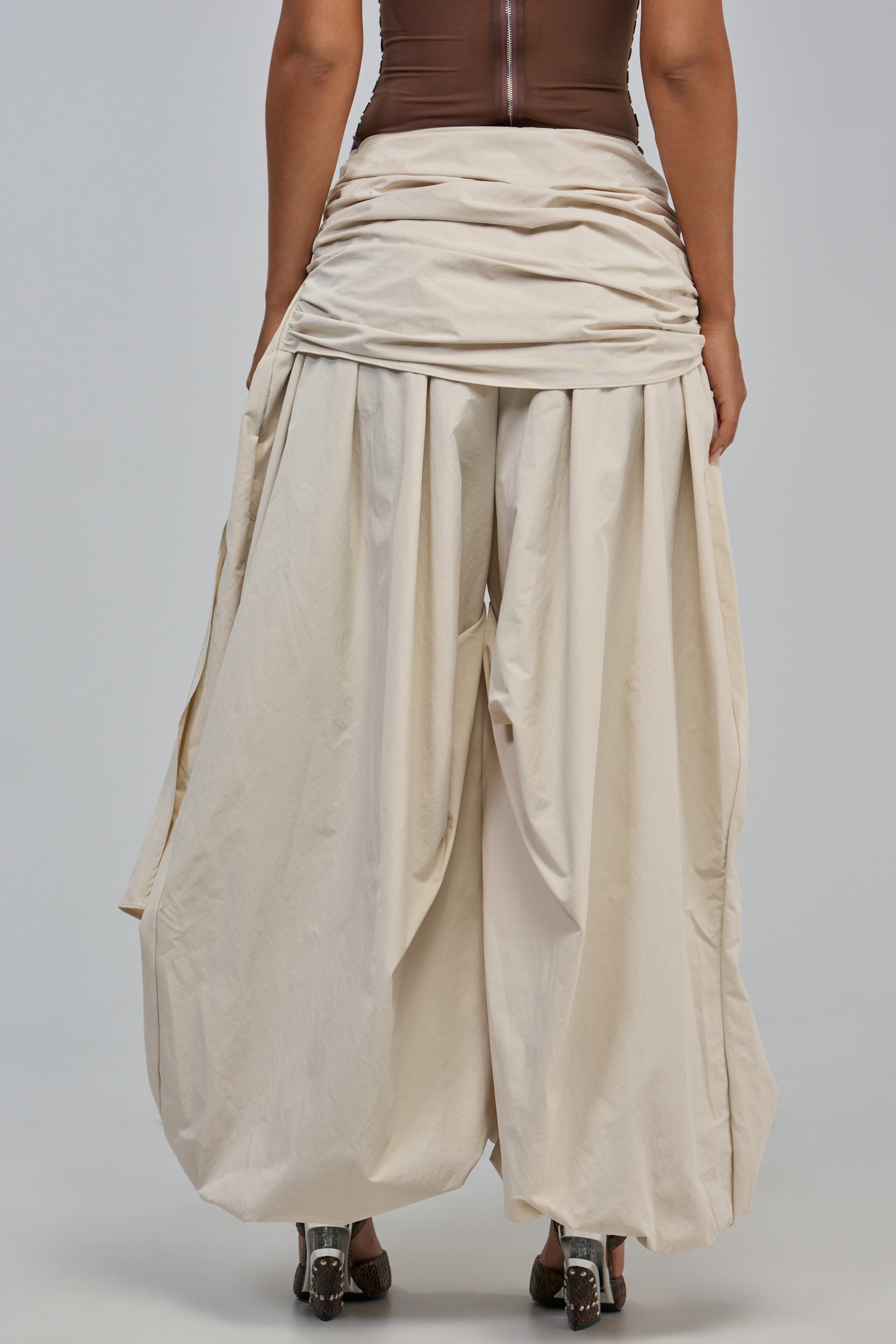A person stands facing away, wearing the NOT MY TYPE TROUSER—a pair of wide, draped cream balloon trousers with a gathered waistband—paired with a brown top. Their shoes are partially visible against a plain, light-colored background.