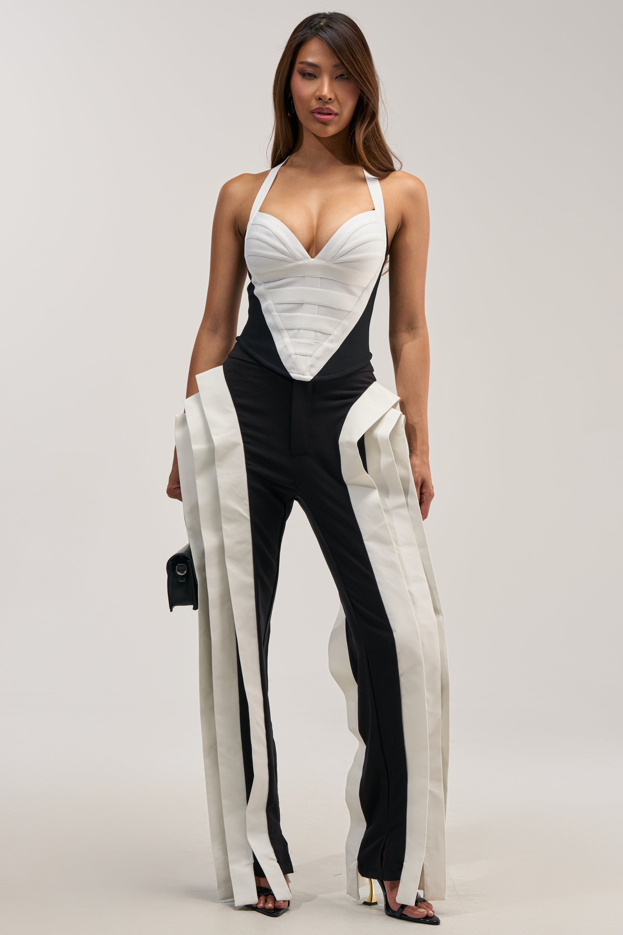 A woman stands confidently against a plain background, holding a small black bag and wearing the INFINITY TROUSER—a black and white business casual statement bottom with dramatic vertical stripes and a structured fit.