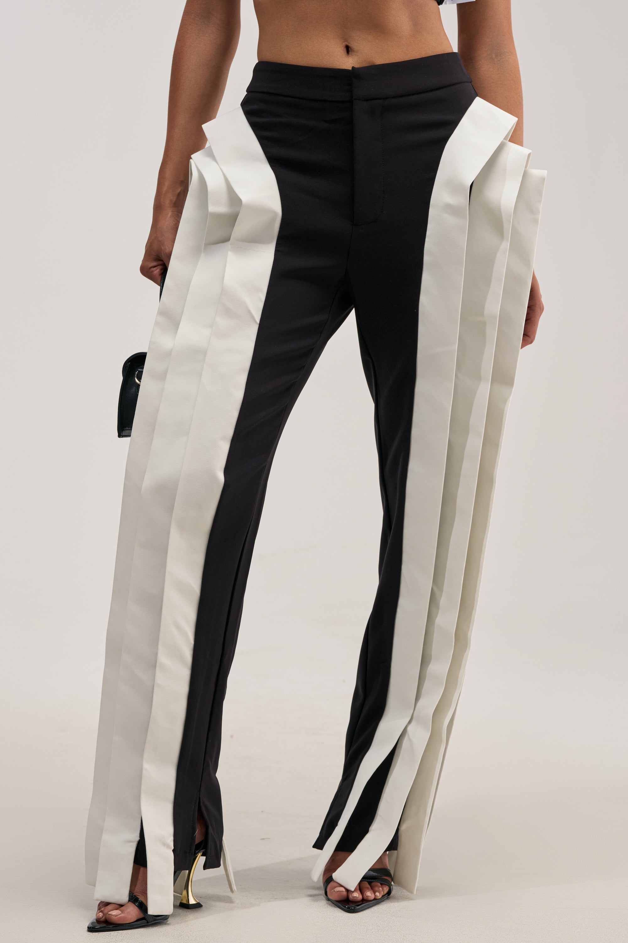 A person wearing the INFINITY TROUSER—a pair of high-waisted black pants featuring wide, vertical white panels that extend to the floor for a statement-making business casual look—paired with open-toe heels. The photo is cropped from mid-torso to feet.