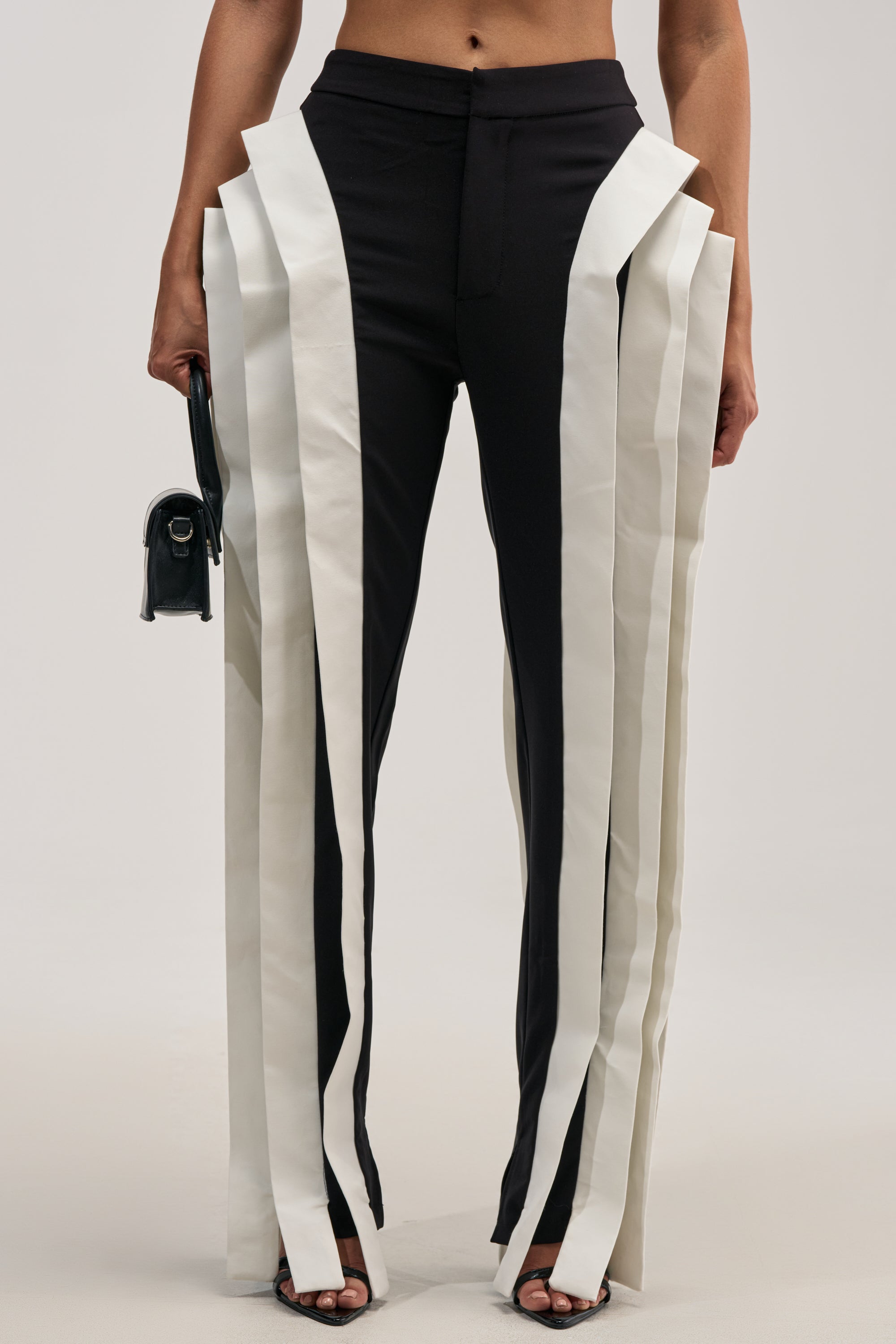 INFINITY TROUSER IN BLACK MULTI: Sleek, tailored straight-leg slim-fit trousers featuring bold white vertical stripe panels that extend outward for an avant-garde look. Styled with a small black bag and black heeled sandals, these statement bottoms offer an editorial edge against a neutral backdrop—ideal for formal occasions when you want to make a striking impression.
