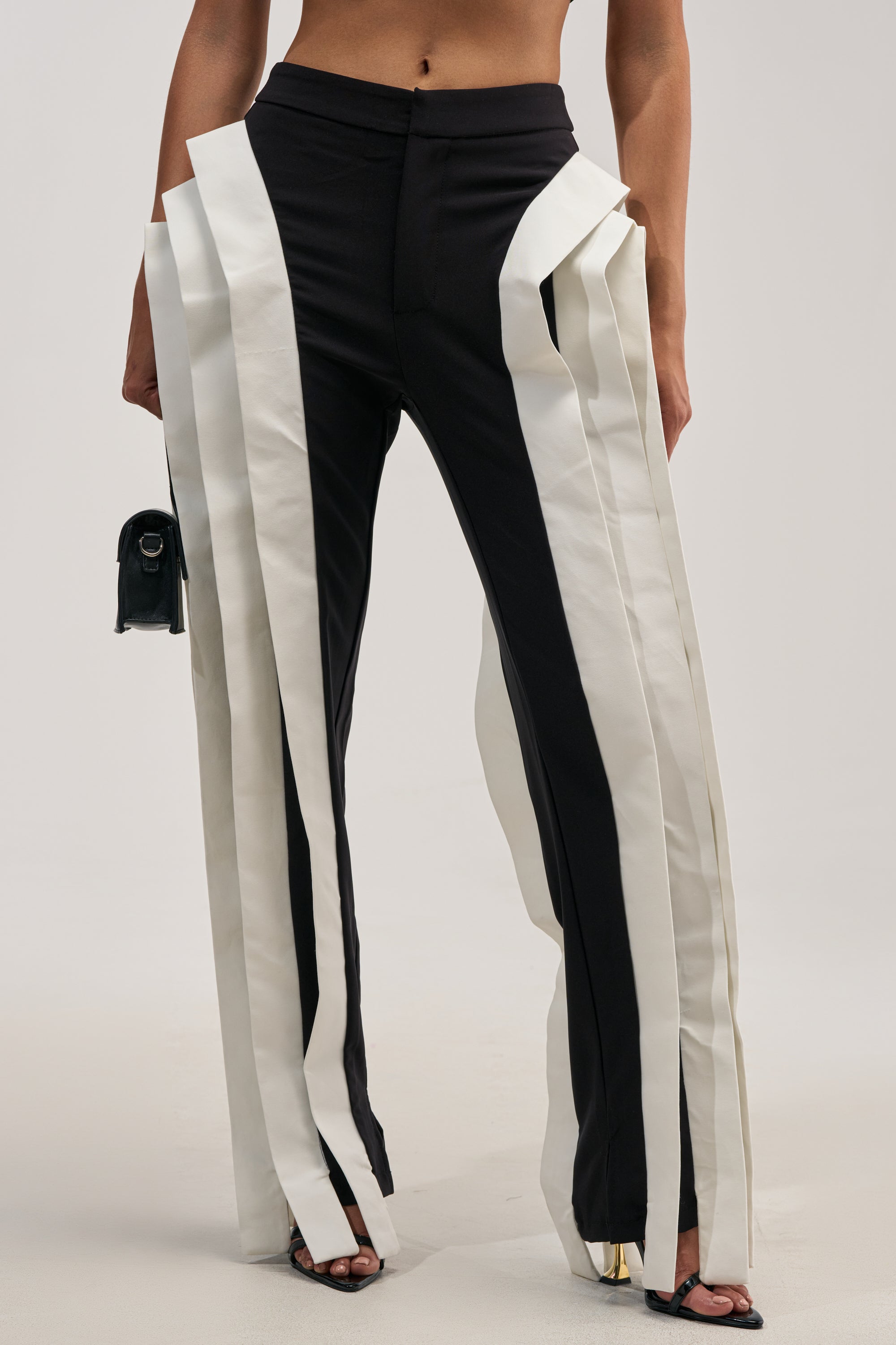 A person wearing the INFINITY TROUSER—high-rise tailored trousers accented with bold vertical white ruffles along each side—paired with a small black handbag and open-toe heeled shoes. Only the lower half of their body is visible.