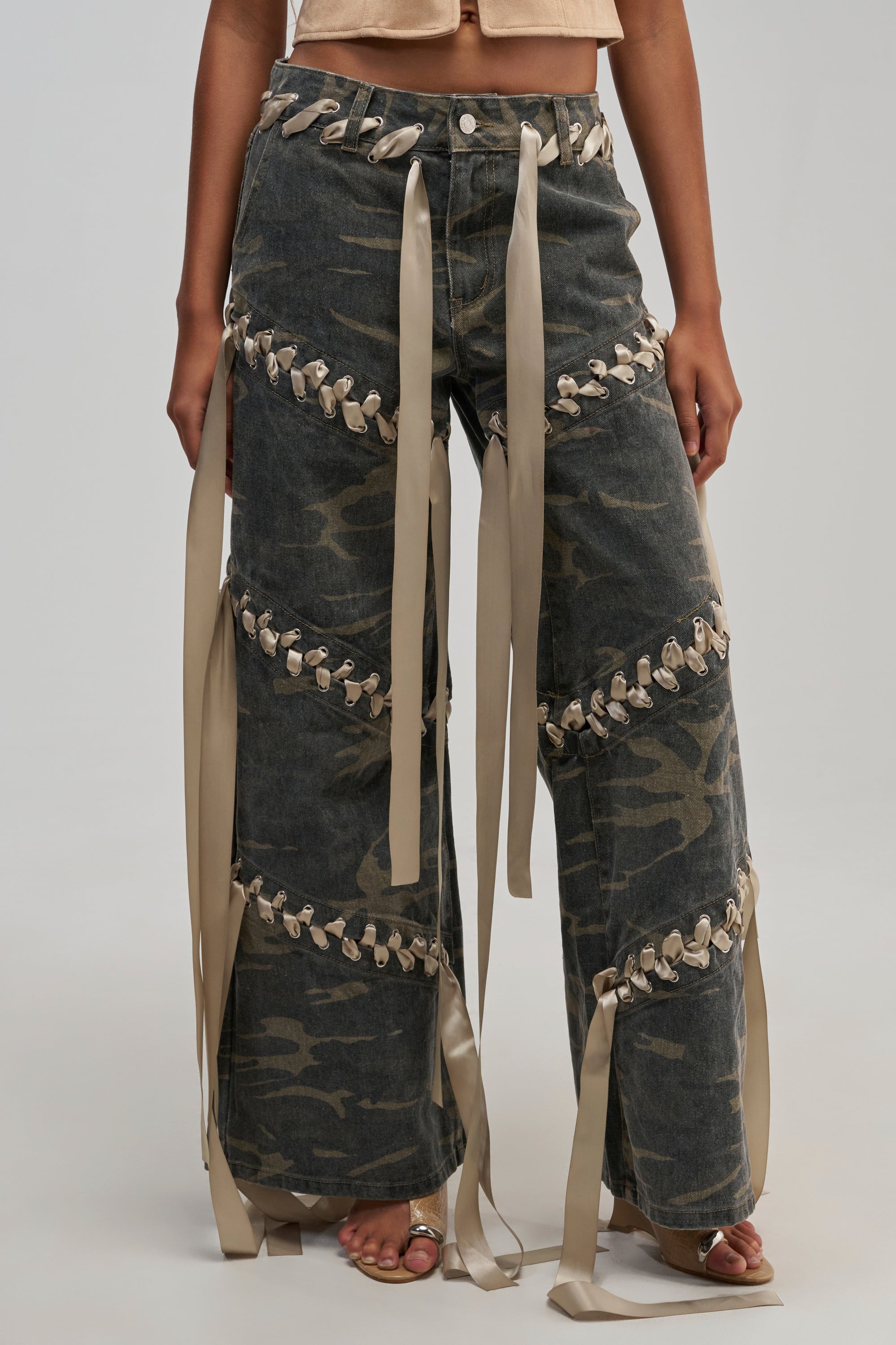A person showcases streetwear style wearing the DOWN FOR SOME FUN CAMO PANT, featuring wide legs, beige ribbon, and lace-up details, standing against a plain background. The utility-chic pants display long, hanging ribbons and are perfectly paired with open-toed shoes. Only the lower body is visible.
