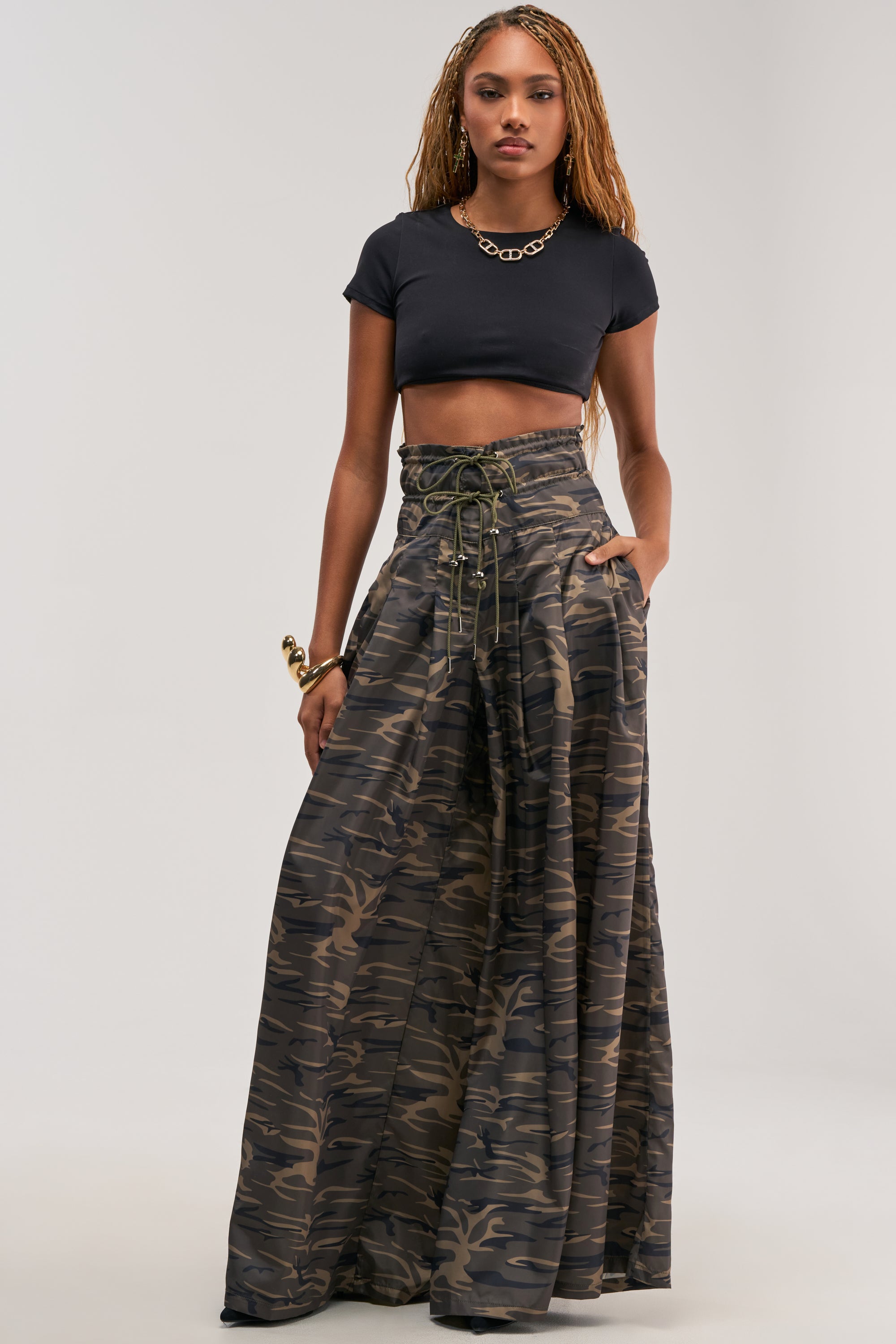 A woman exudes confidence in the “DANCE THE NIGHT AWAY CAMO PALAZZO PANT,” pairing these wide-leg camouflage pants with a black crop top and high-waisted streetwear. Her look is finished with a chunky gold necklace and bracelet, as she stands with hands in pockets against a plain background.