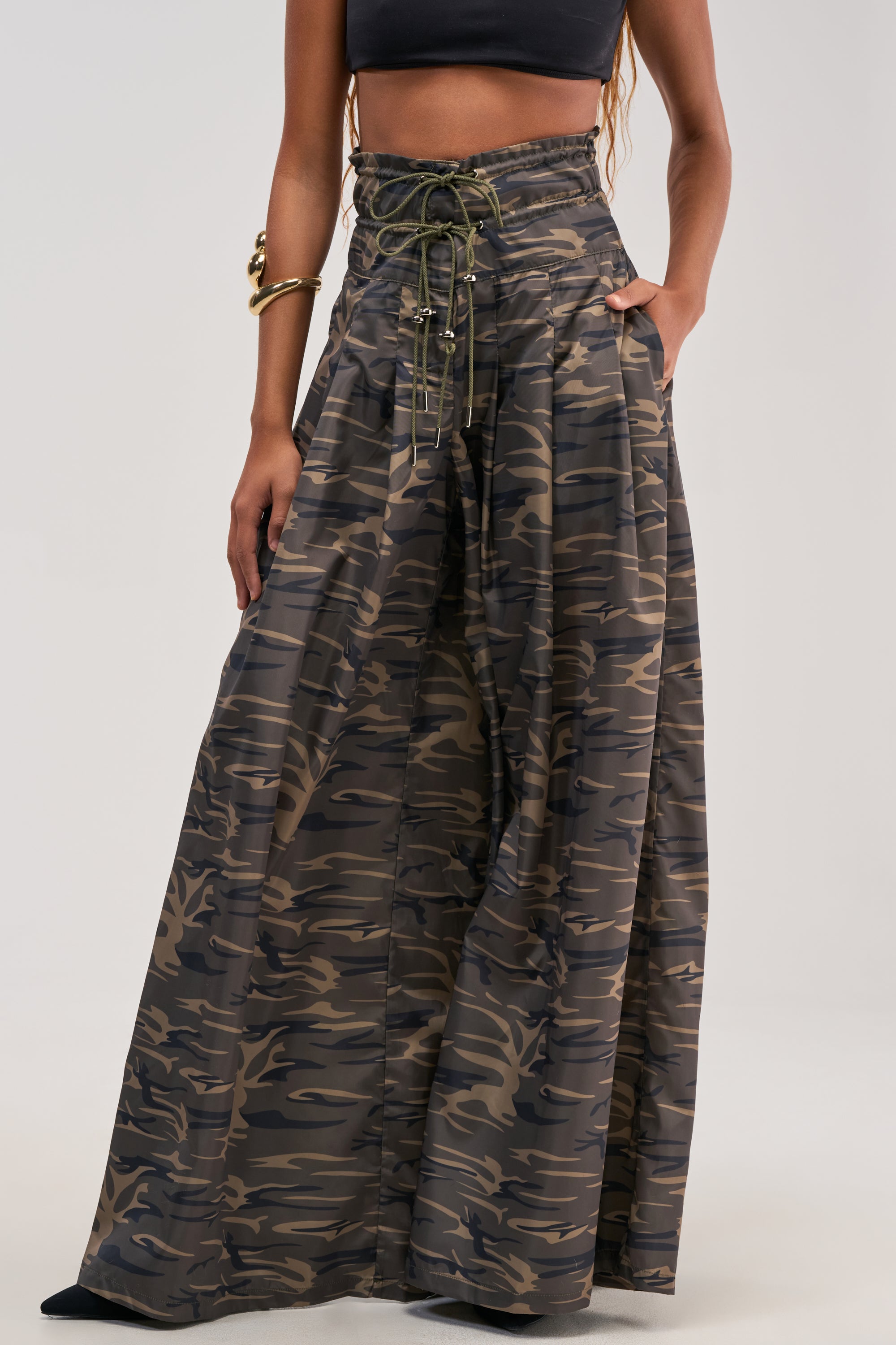 Rocking the DANCE THE NIGHT AWAY CAMO PALAZZO PANT with its high-waisted fit and lace-up front, paired effortlessly with a black crop top and a gold bracelet. One hand is tucked in the pocket, while the shot is cropped above the shoulders to highlight this bold streetwear-inspired look.