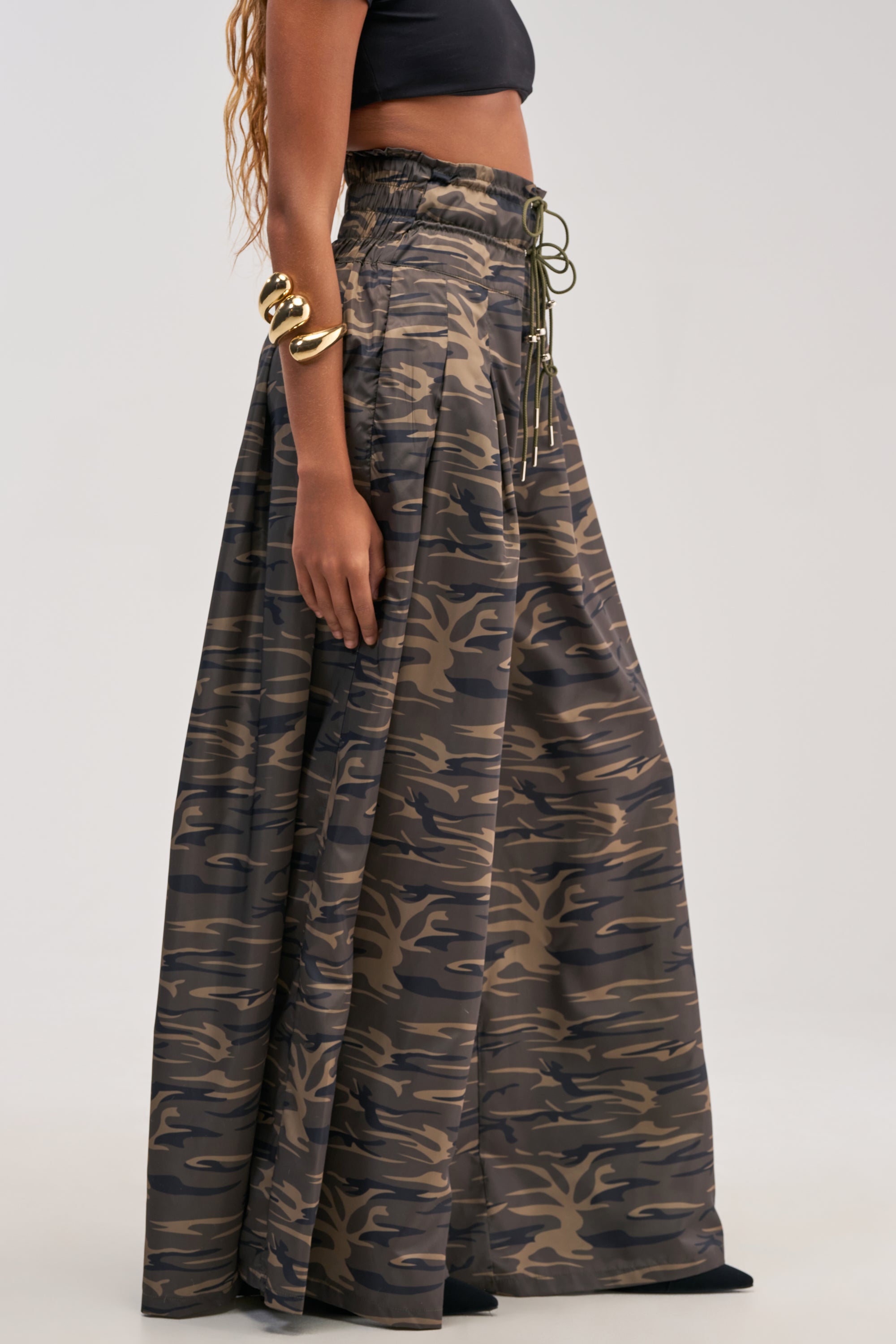 Dressed in the DANCE THE NIGHT AWAY CAMO PALAZZO PANT, featuring a high-waisted fit and ruched drawstring waist, paired with a black crop top, this individual radiates effortless streetwear style. Chunky gold bracelets adorn their arm against a simple, light-colored backdrop.