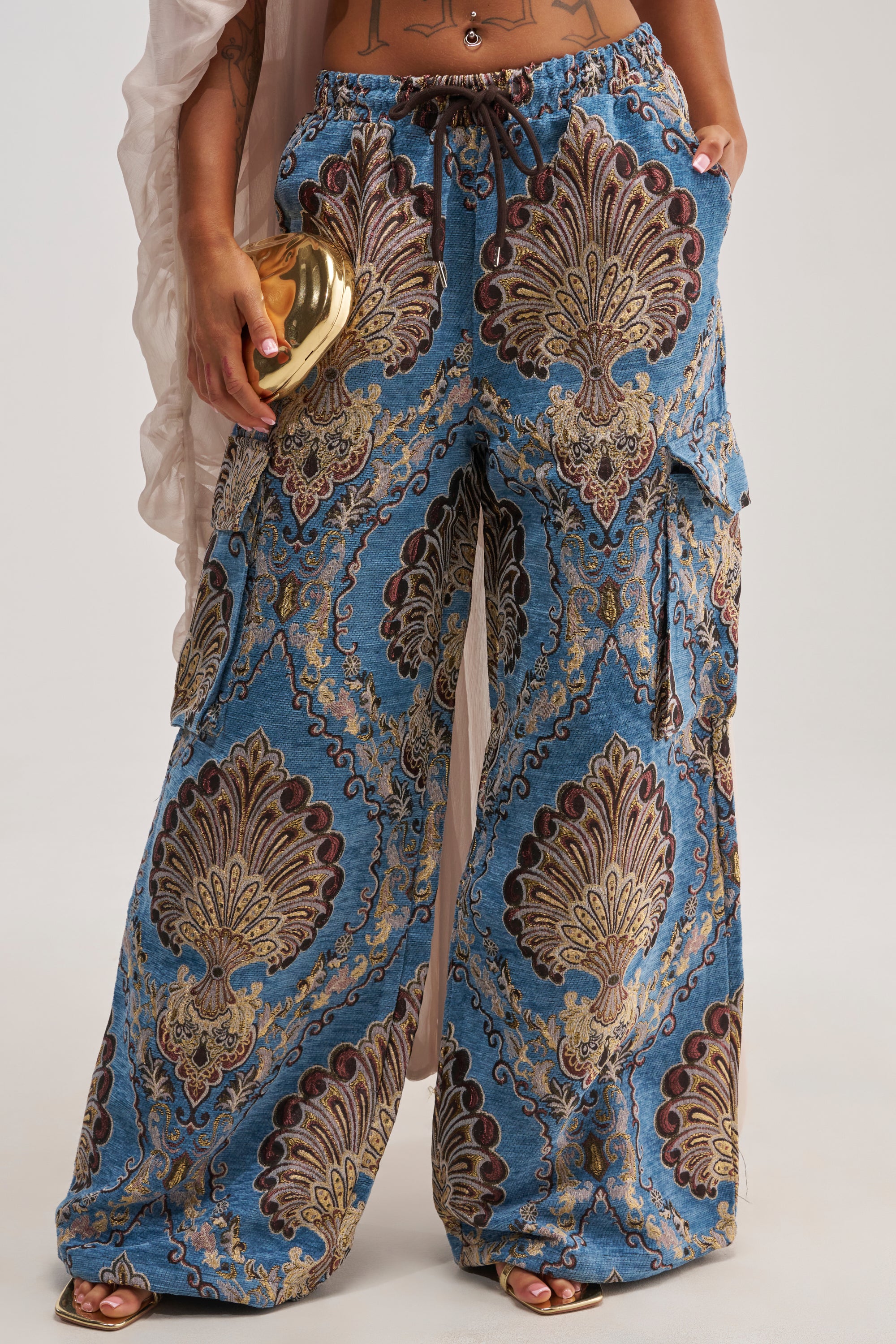 A person is shown from the waist down wearing the DUOMO CARGO JOGGER, featuring wide-leg blue patterned pants with an ornate gold baroque print and maroon accents. They are holding a small shiny gold clutch and wearing open-toe sandals, set against a plain white background.