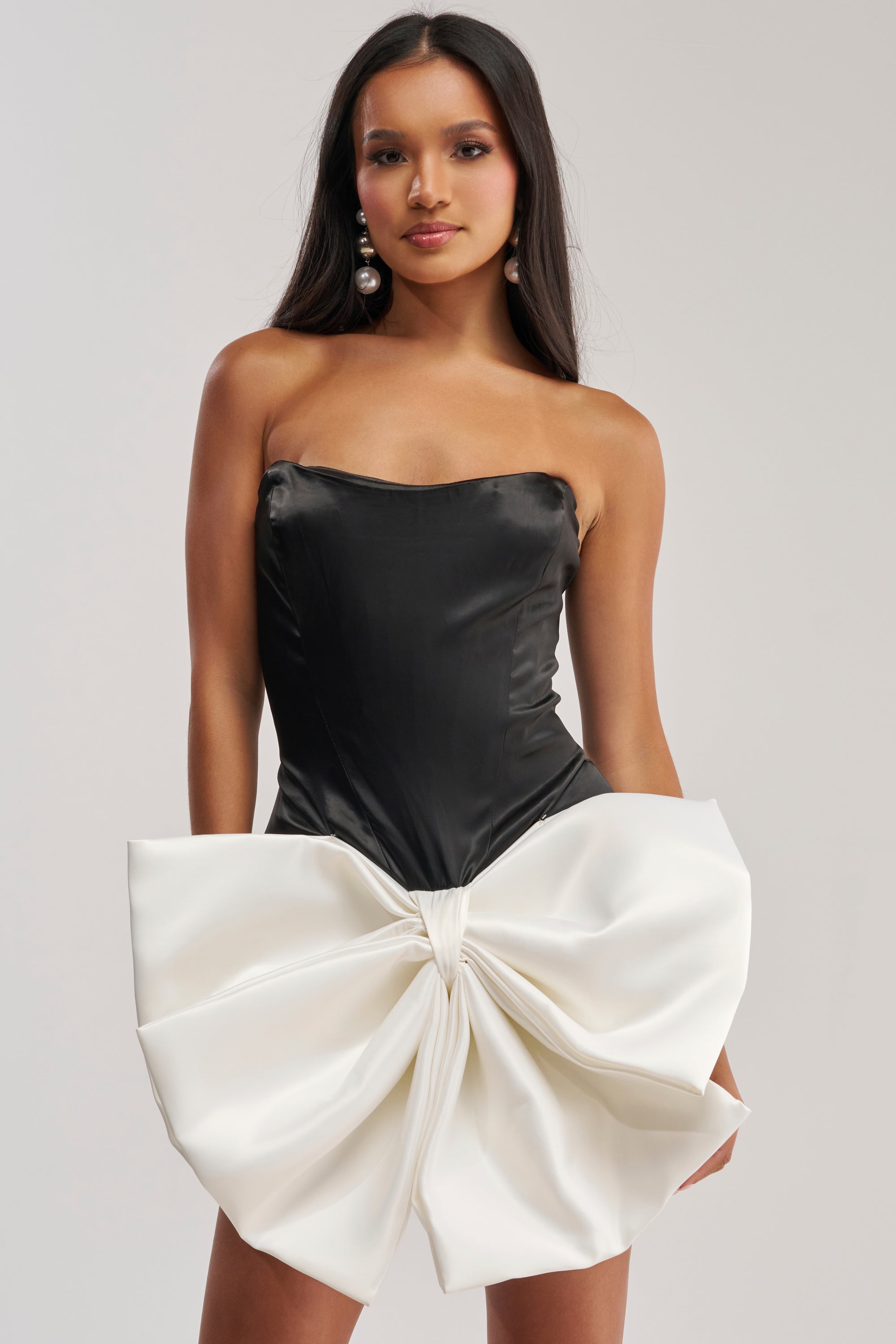 A woman models the SITTING PRETTY BOW MINI DRESS, a strapless black satin mini with a large sculpted white bow at the skirt. She accessorizes with statement pearl earrings and poses against a simple light background.