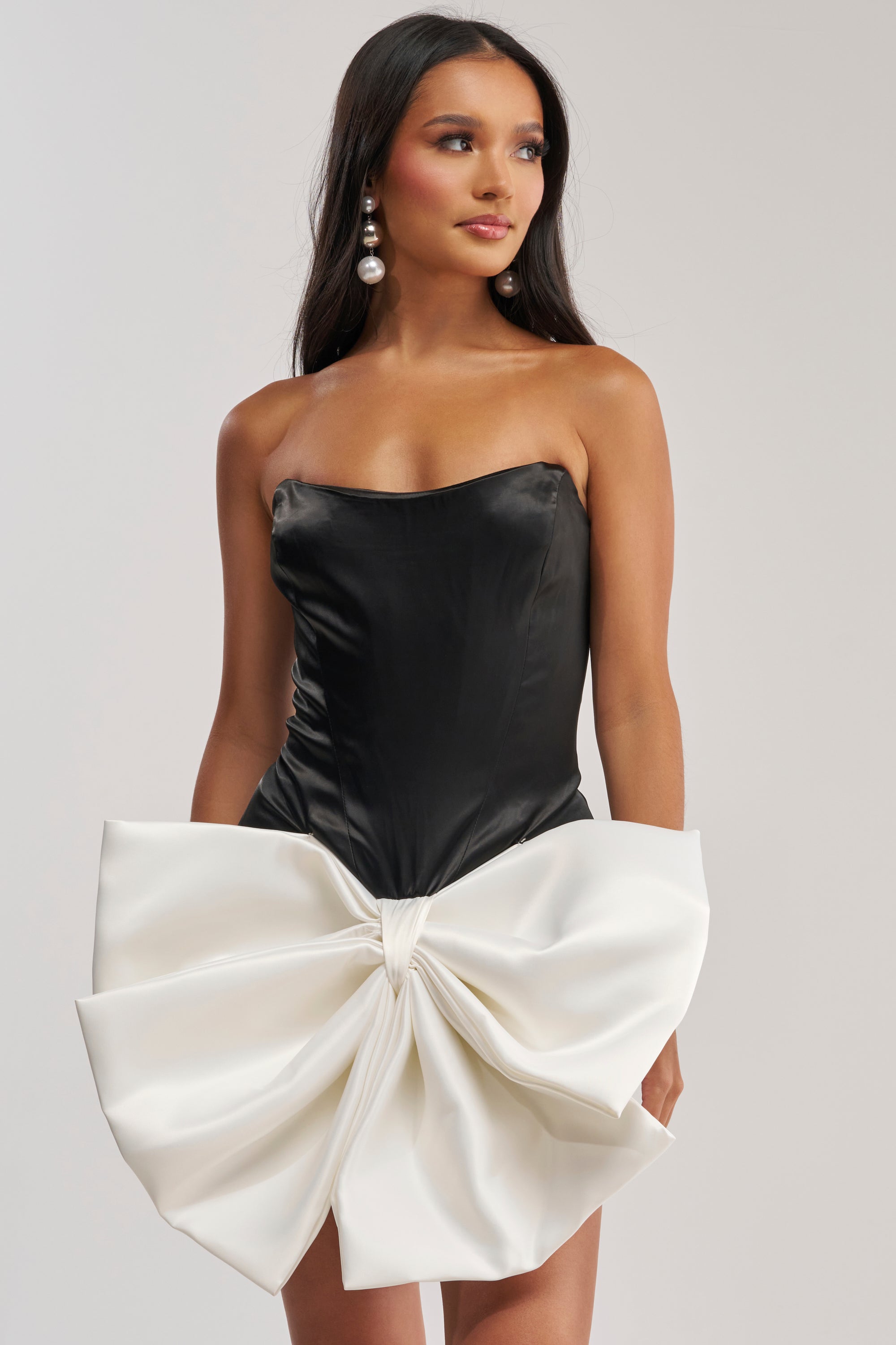 A woman with long dark hair and pearl earrings stands against a plain background wearing the SITTING PRETTY BOW MINI DRESS, a strapless black satin mini dress featuring a large sculpted white bow at the waist.
