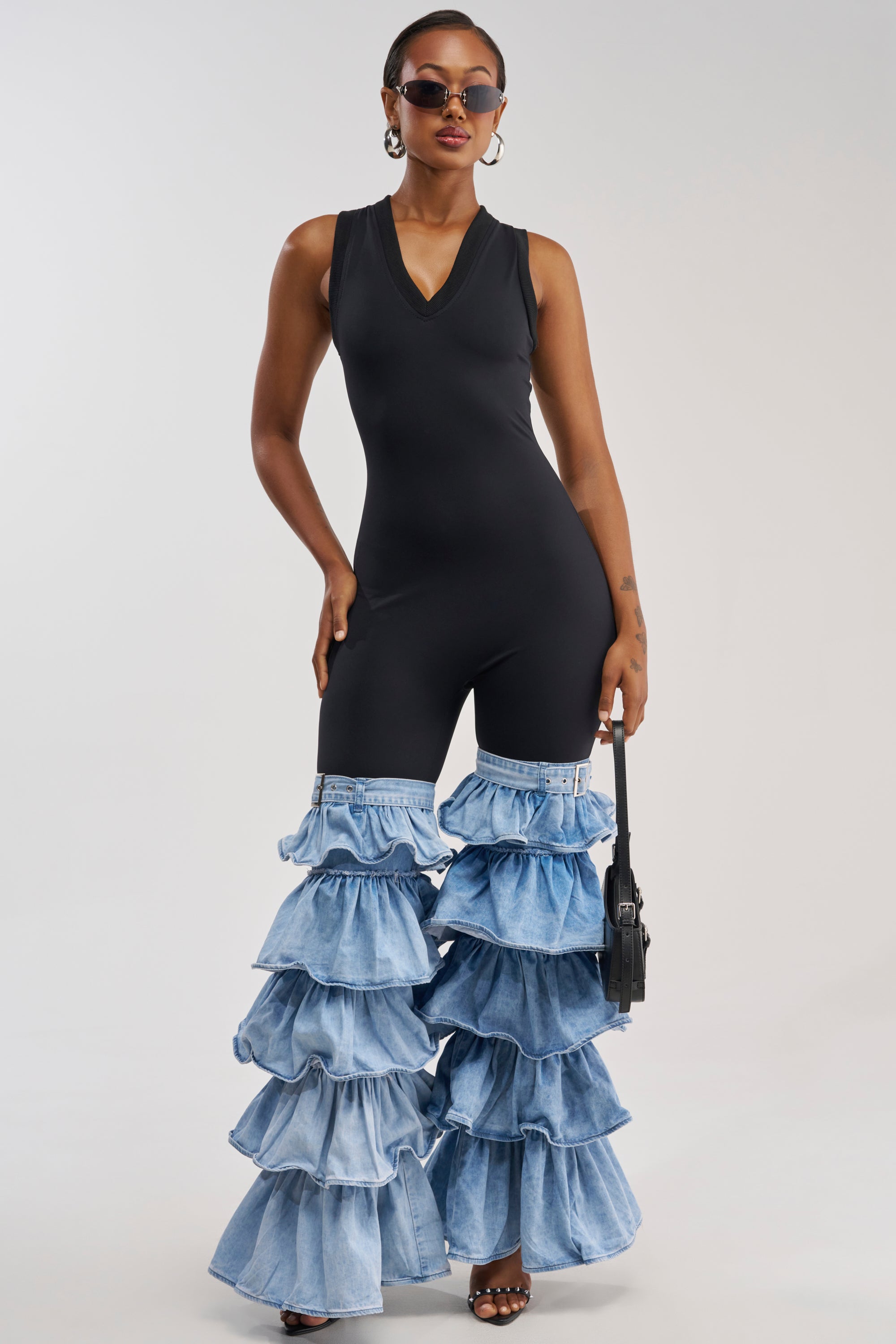 A woman models the ELEVATED BASICS BODYCON JUMPSUIT WITH RUFFLE DENIM LEG—a sleeveless black jumpsuit featuring tiered denim ruffle bell bottoms. She accessorizes with sunglasses, hoop earrings, heels, and a small black purse while standing against a plain light background.