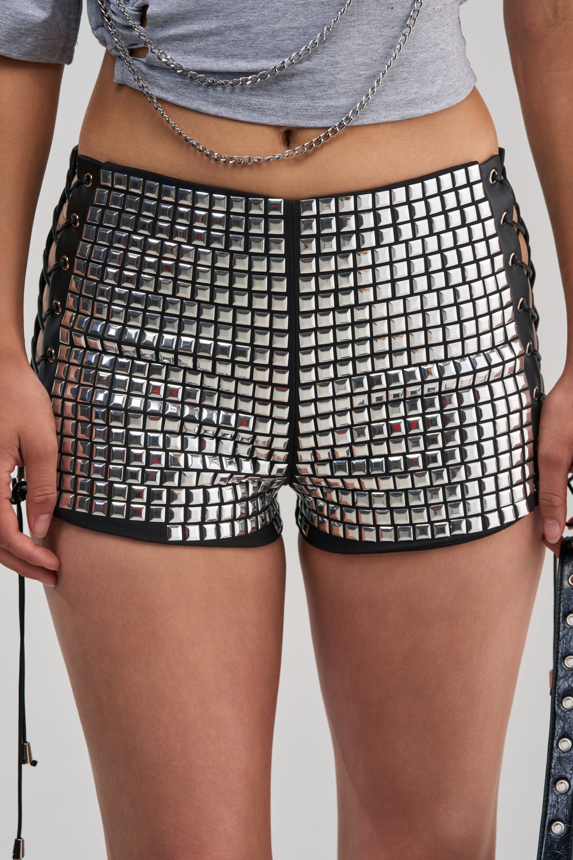Wearing the PARTY TIME HOT SHORT—a pair of high-rise, shiny metallic shorts embellished with square studs—paired with a cropped gray top accented by chains, as seen in a photo capturing the look from mid-torso to mid-thigh.