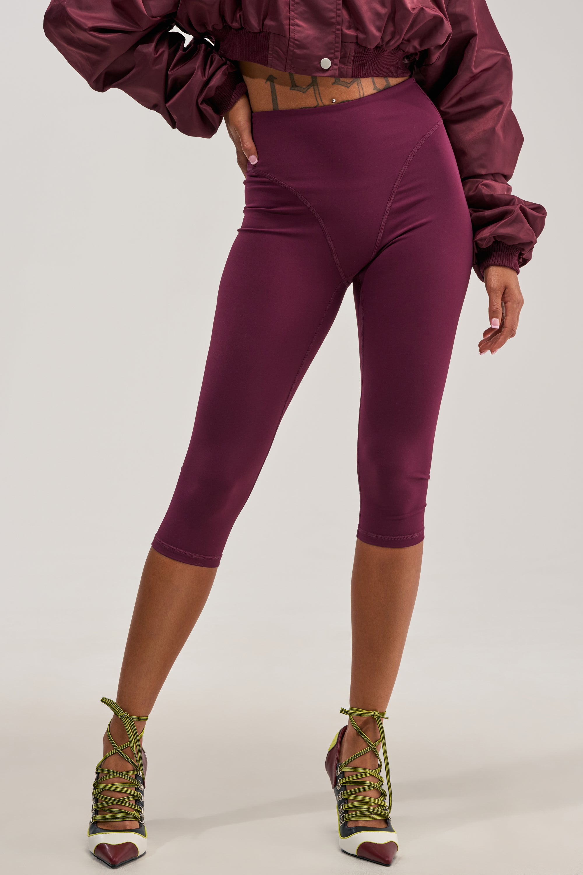 A person wears the EASY ENOUGH CAPRI—high-waisted maroon capri leggings featuring a high-rise waist—paired with a matching cropped jacket and green lace-up heels. The face is not shown, and the plain light gray background highlights the chic, sportswear-inspired ensemble.