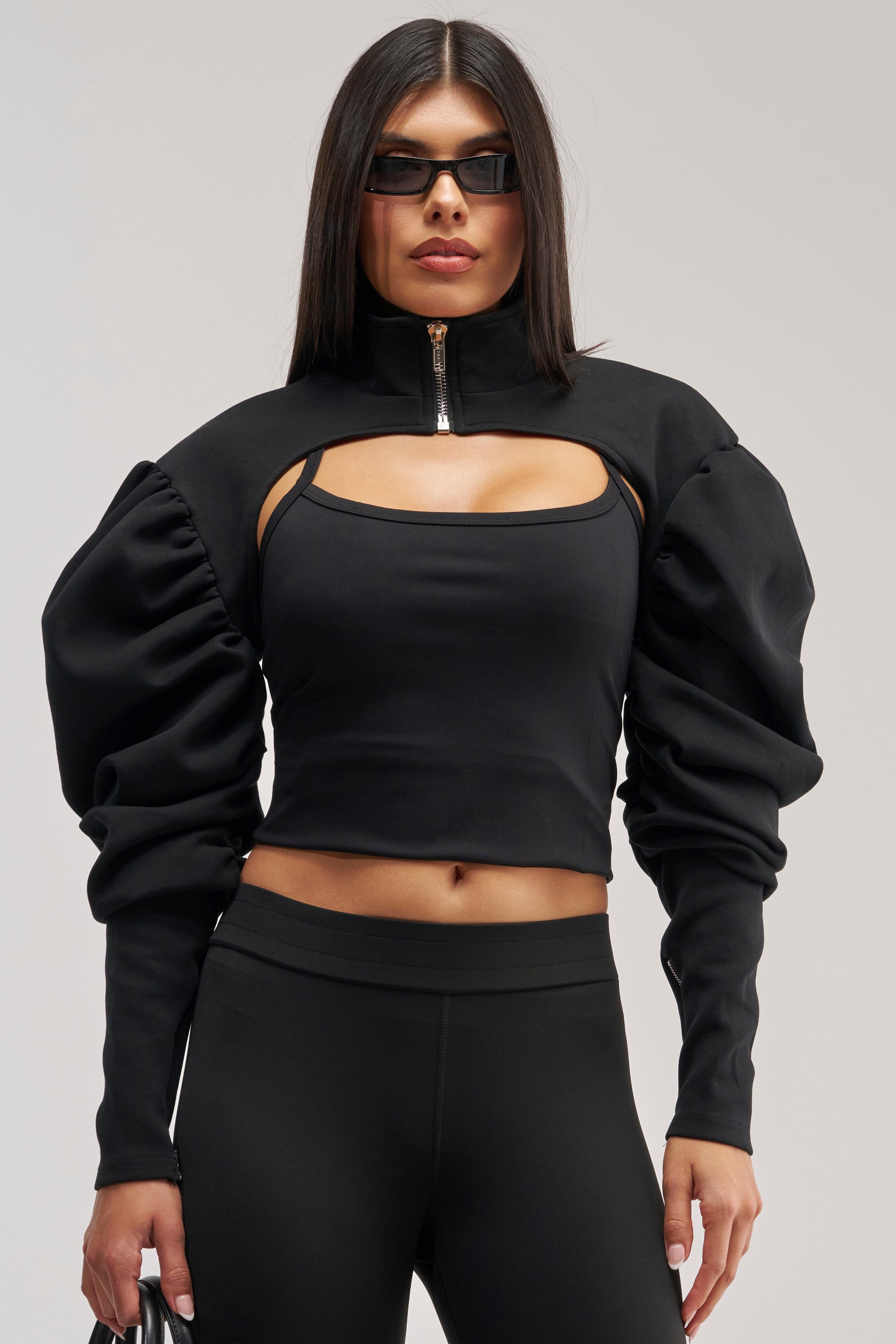 A woman wearing black sunglasses, a black top styled as the SCARLETT RUCHED SLEEVE BOLERO with front cut-out and puffed long sleeves, paired with black leggings, stands against a neutral background.
