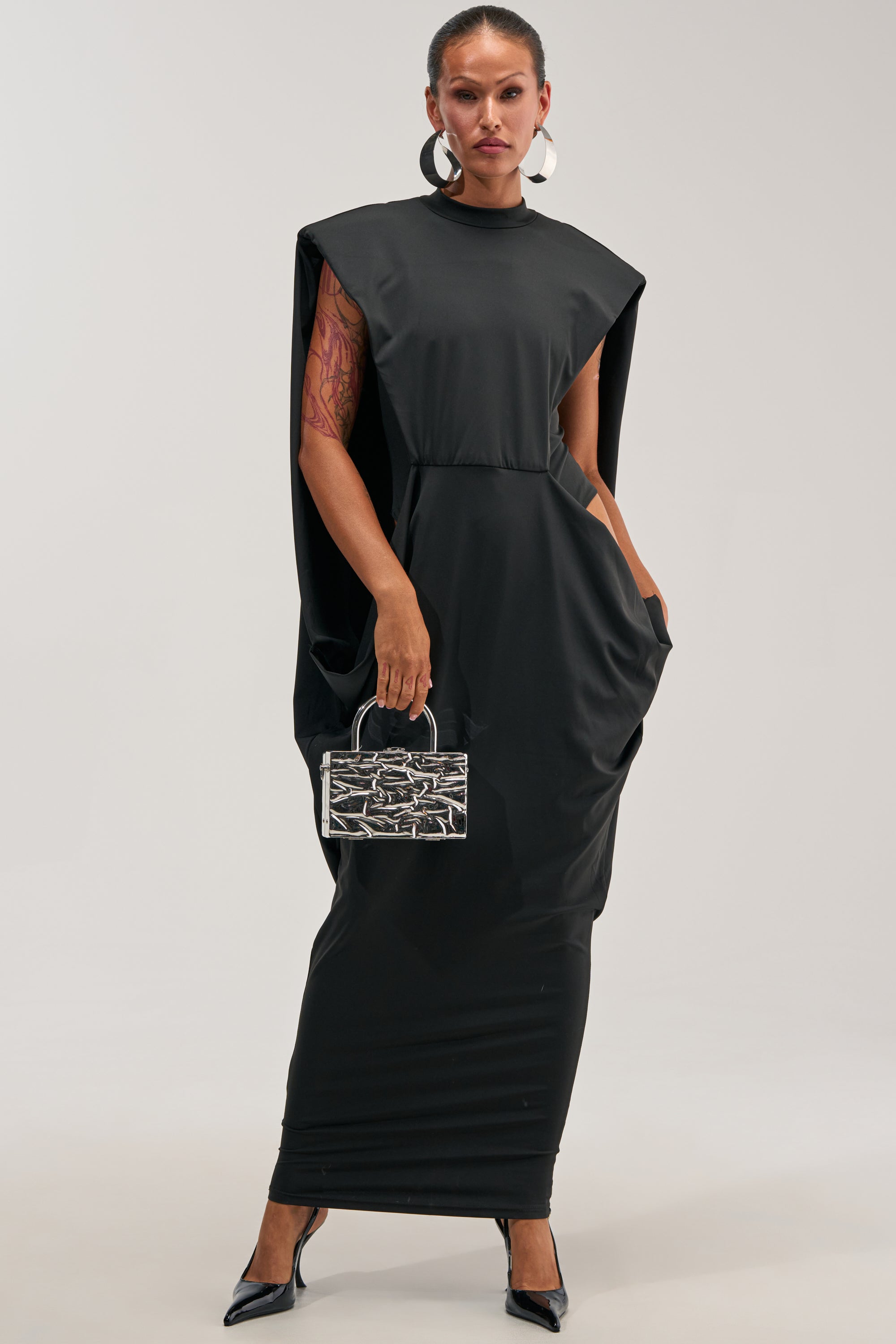 Wearing the SWEET TALKER CAPE MAXI DRESS—a long, structured black maxi dress featuring elegant cape sleeves—a woman holds a small silver textured handbag and accessorizes with black heels and large hoop earrings. Poised confidently against a plain, light background, she embodies the ideal evening dress or sophisticated wedding guest look.
