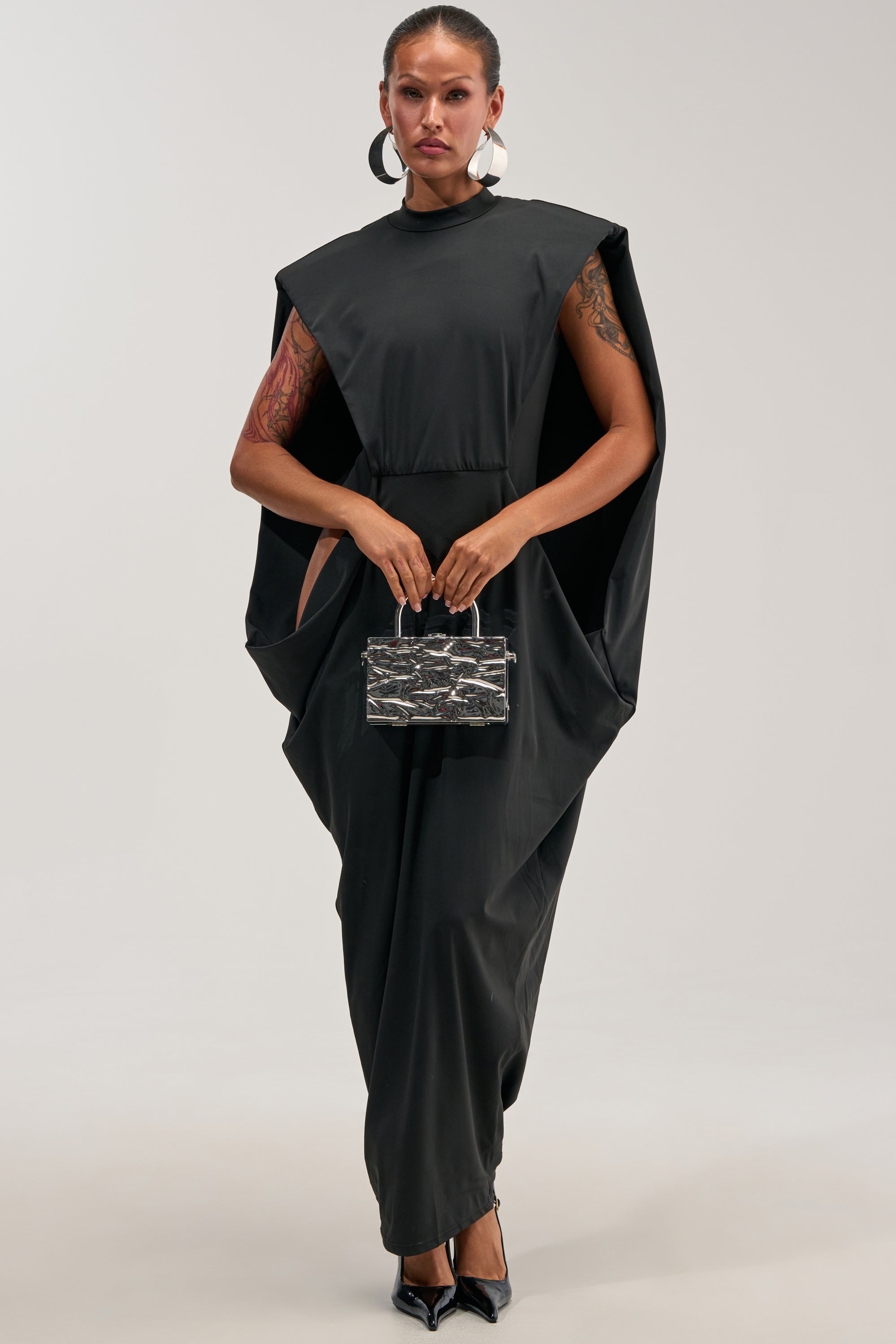 A woman stands confidently in the SWEET TALKER CAPE MAXI DRESS, a dramatic black maxi dress with a cape detail, accessorized with a small clear handbag, black heels, oversized hoop earrings, and showcasing her arm tattoos. Set against a plain light background, her look is effortlessly bold—ideal for evening events or as an unforgettable wedding guest ensemble.