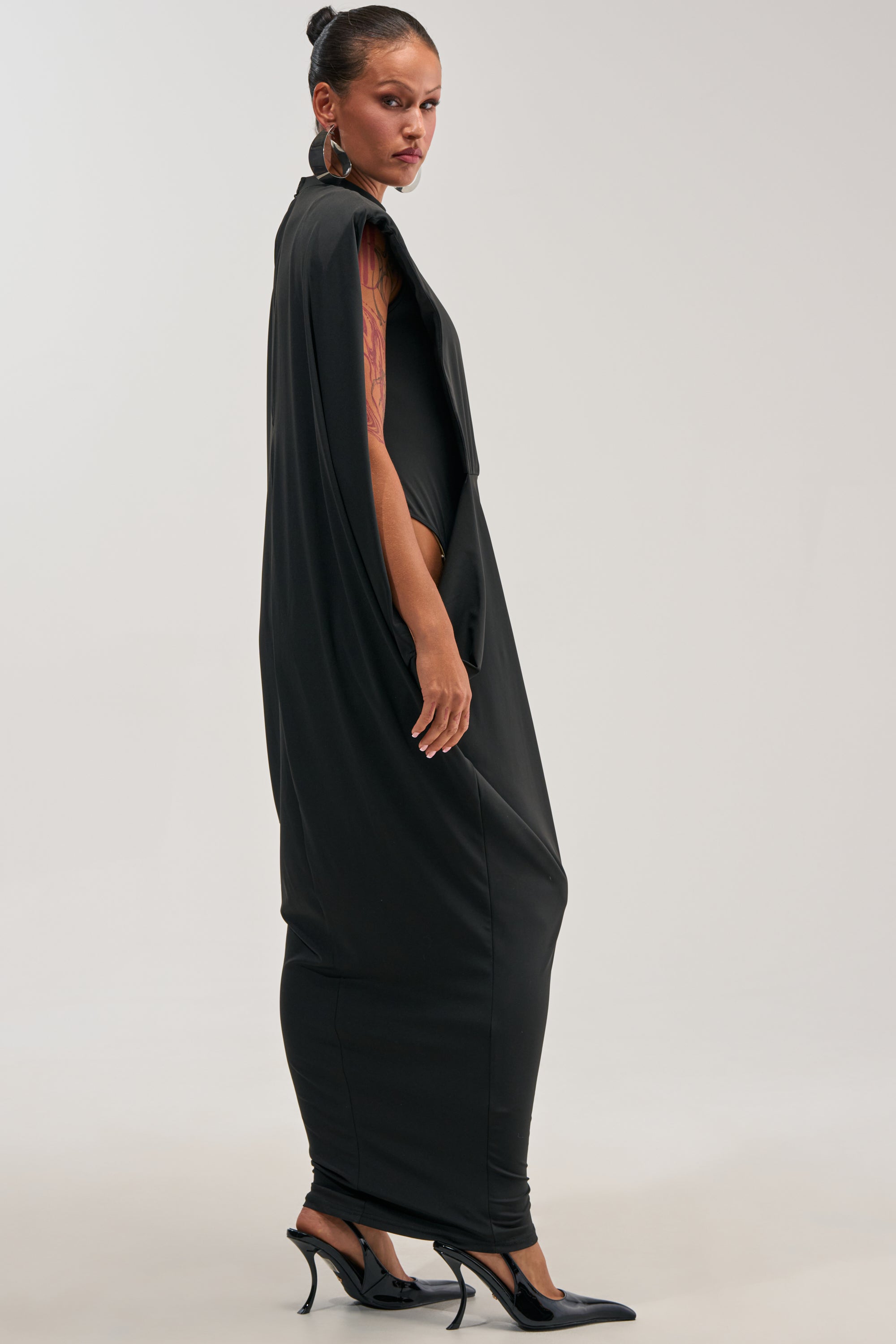 A woman stands in profile wearing the SWEET TALKER CAPE MAXI DRESS, a striking black maxi dress featuring dramatic cape-like sleeves, paired with black heels. Her hair is styled in a sleek bun, showcasing a tattoo on her upper arm. Set against a simple, light background, this elegant evening dress is the perfect choice for special occasions or as a chic option for wedding guests.