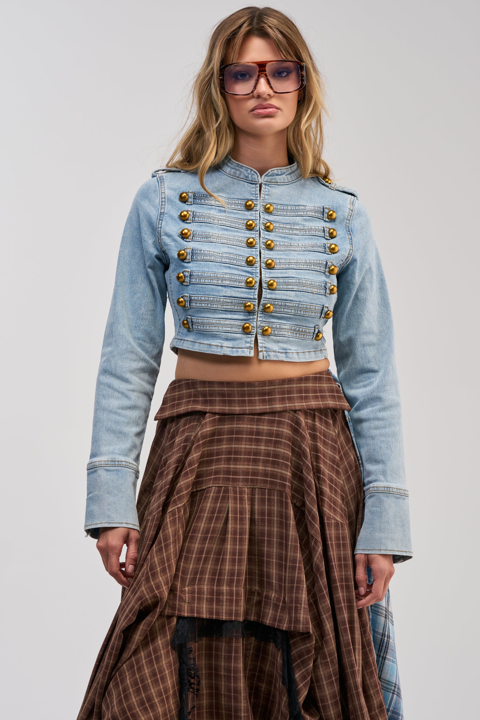 Wearing the POWER MOVE NAPOLEON JACKET IN DENIM—a light blue, military-inspired piece with gold buttons—a person stands against a plain background, styled with a layered brown plaid skirt and large square sunglasses.