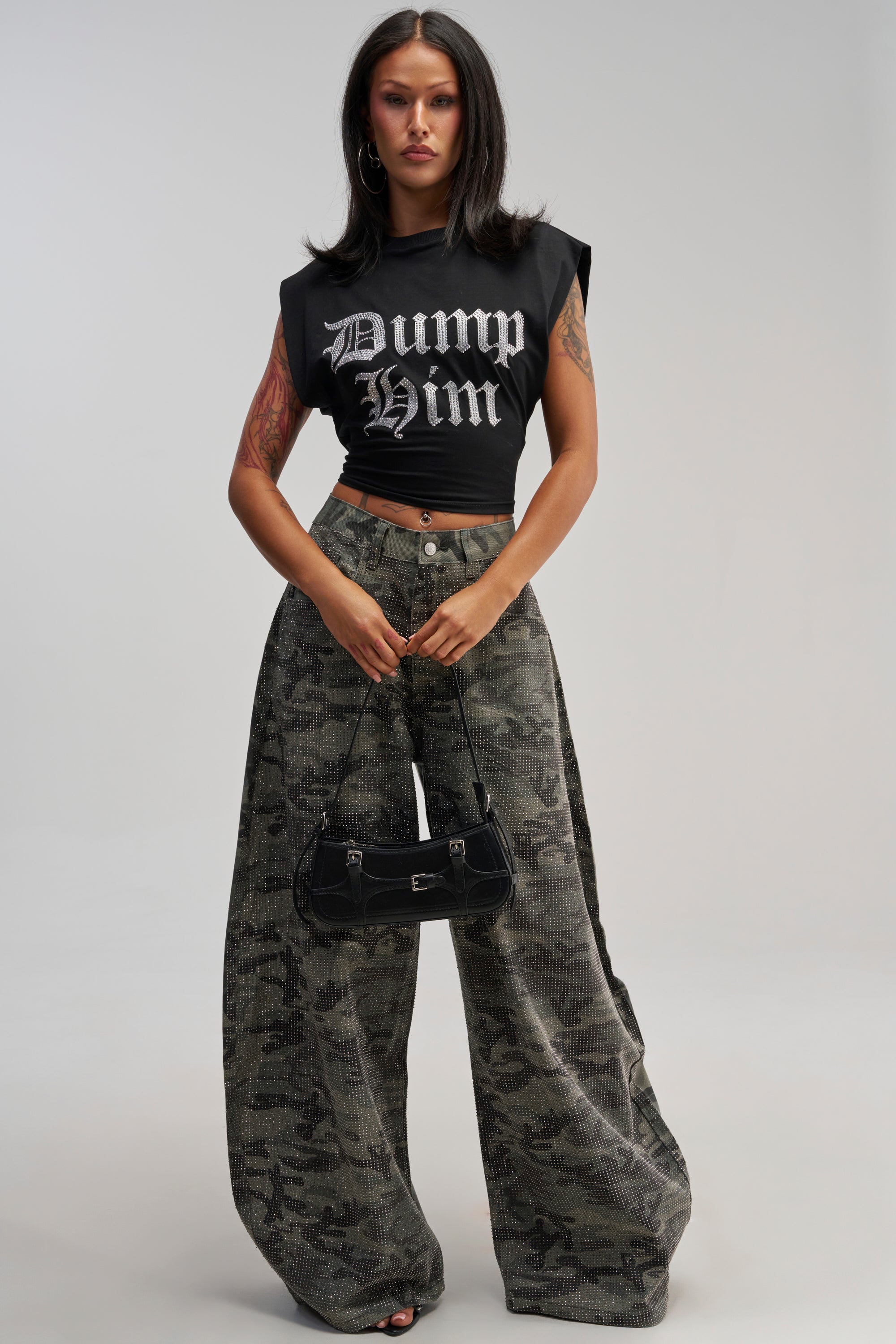 A woman with straight dark hair wears a sleeveless black “Dump Him” crop top, CAMO FOR DAYS BARREL PANT, and holds a small black handbag, standing against a plain light background.
