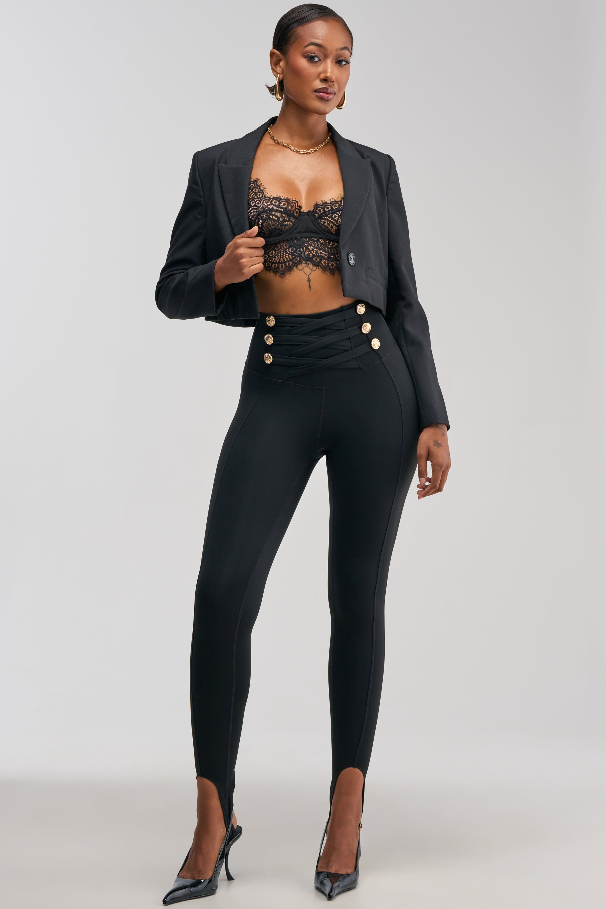 A woman stands confidently against a plain background, wearing the COME AND BUY IT STIRRUP LEGGING paired with a black cropped blazer, lace bralette, and black heels.